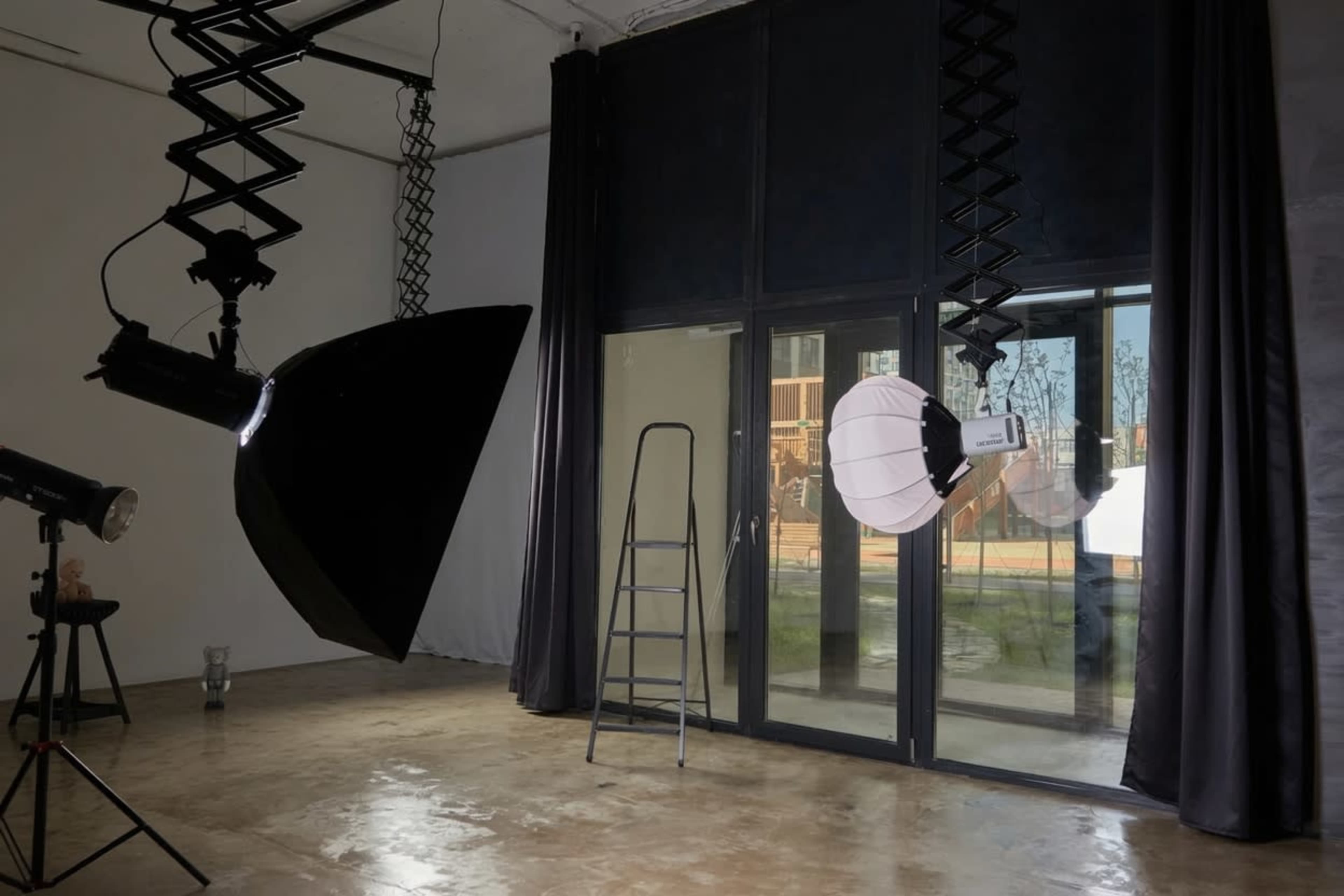 The image shows a photography studio with two hanging light fixtures and a ladder in front of large windows.