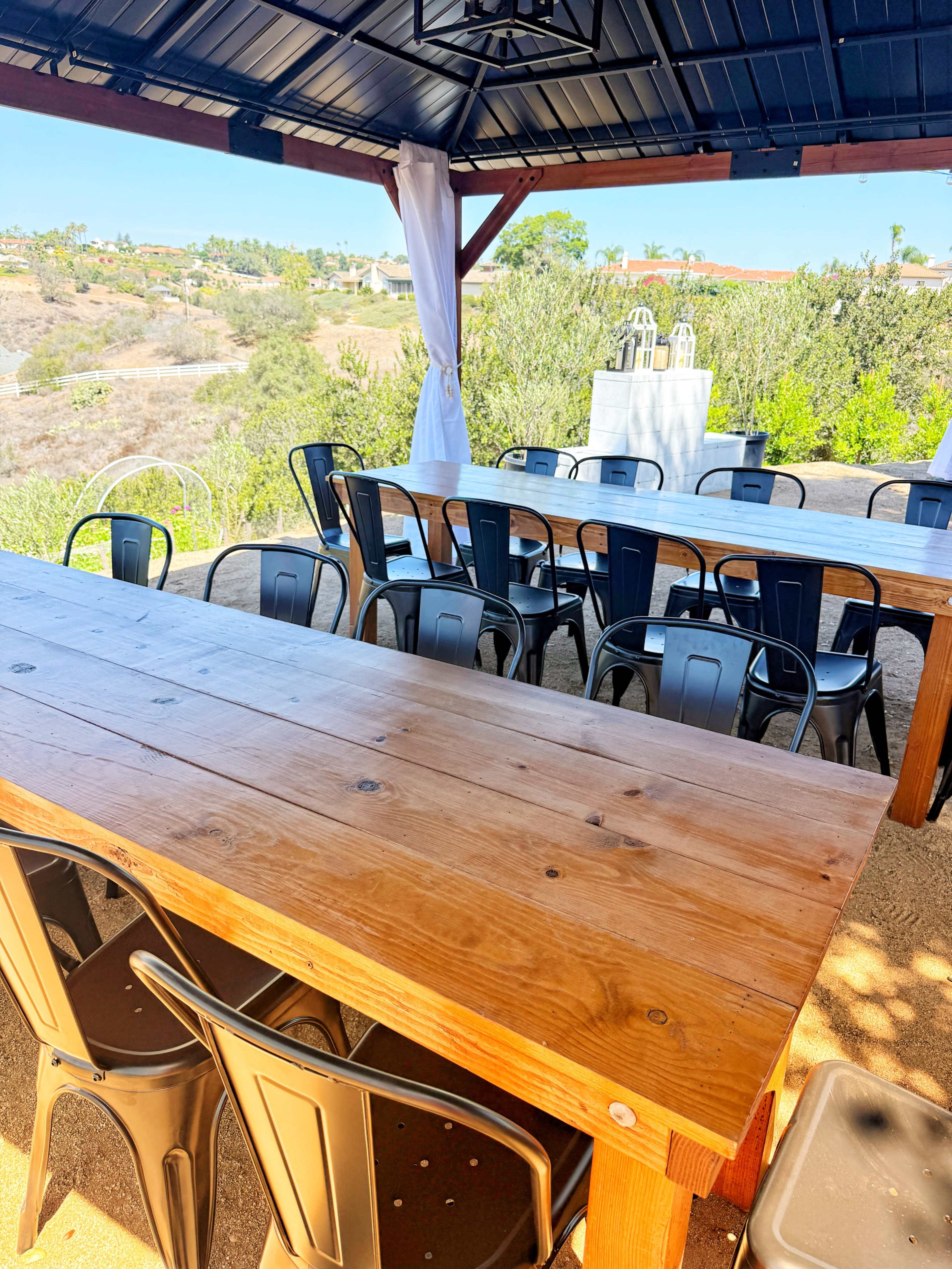 A spacious outdoor dining area features long wooden tables with black metal chairs under a gazebo, surrounded by greenery and an empty landscape.