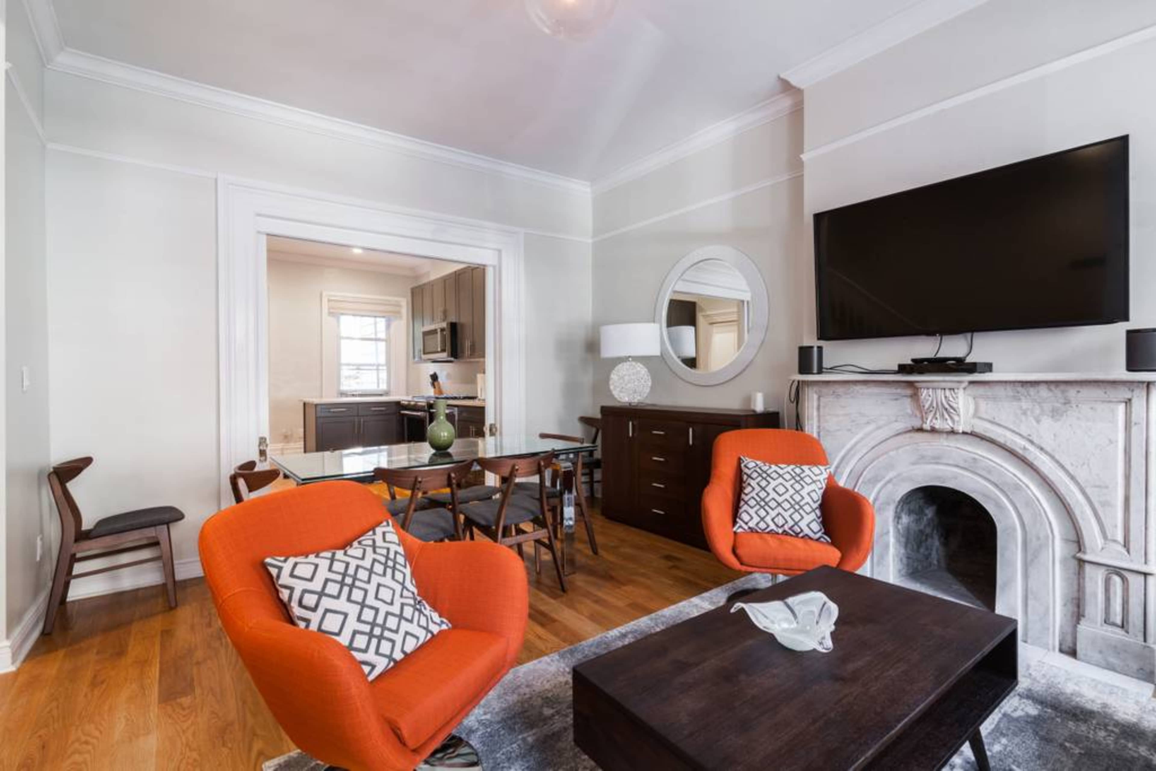 A modern living room featuring orange armchairs, a dark wooden coffee table, a marble fireplace, and a television mounted on the wall, with an adjoining kitchen visible in the background.