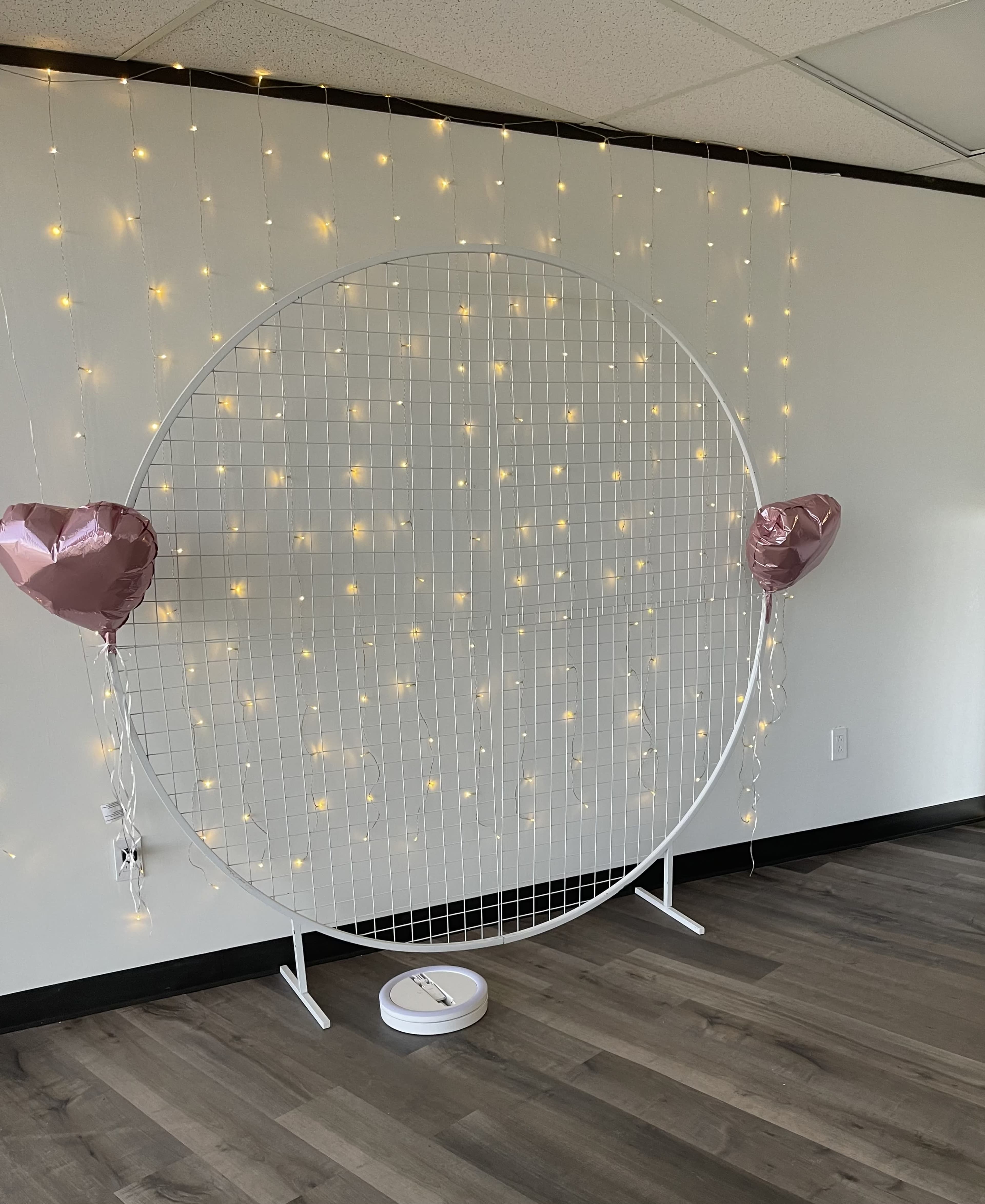 A large circular frame with a grid design is set against a wall adorned with string lights, and two heart-shaped balloons are attached to the frame.
