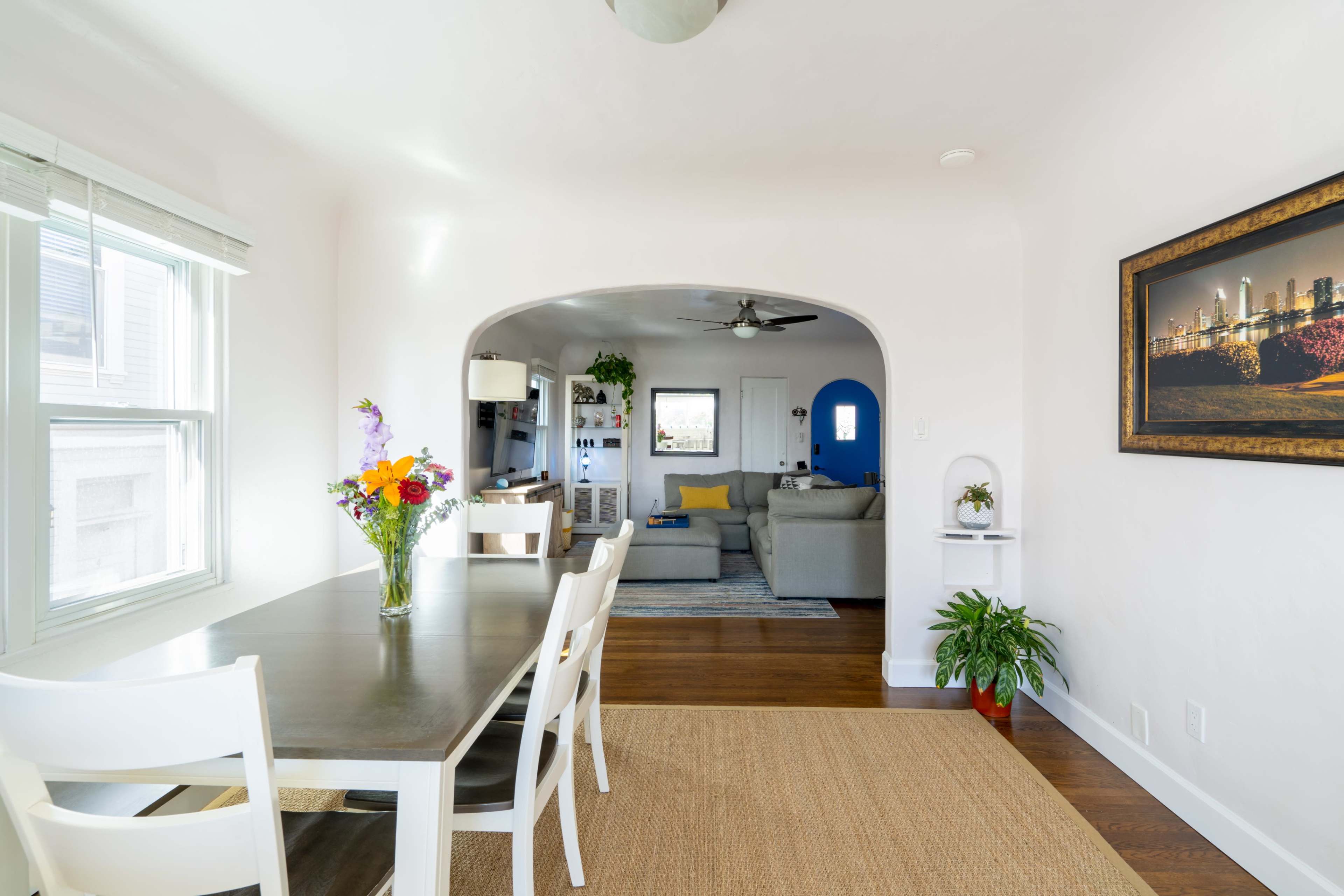 A dining area featuring a long table with flowers, leading into a living room with a couch and blue accent wall.