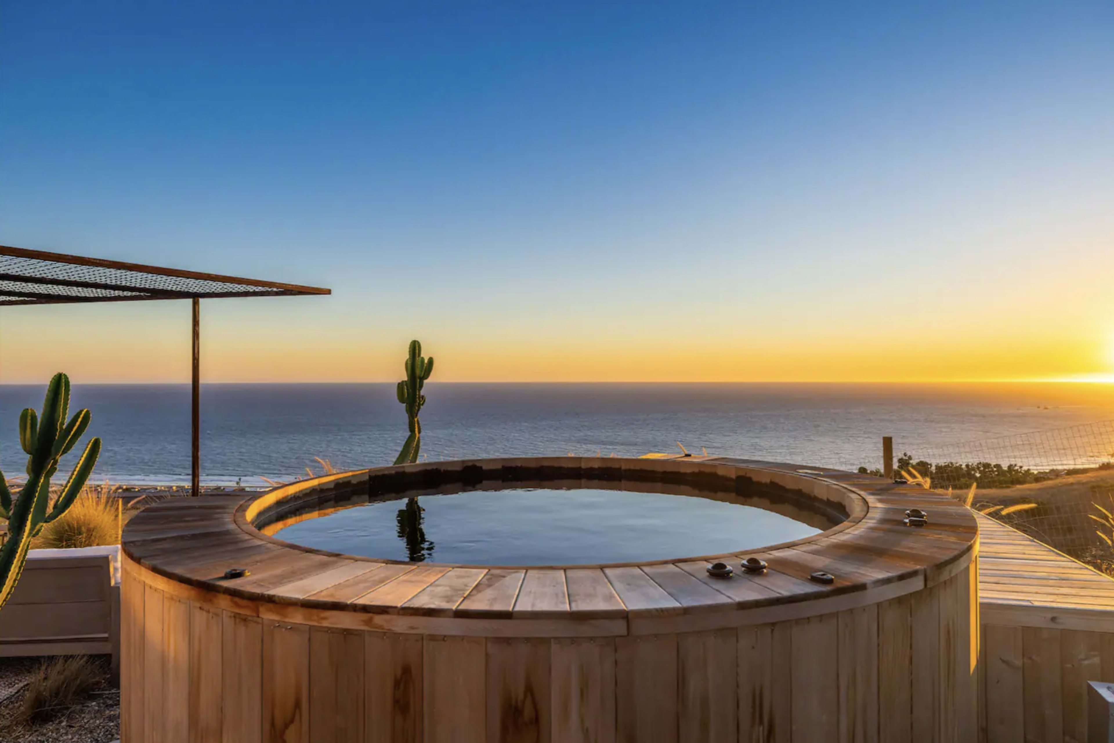 The image shows a wooden hot tub overlooking the ocean at sunset, with a clear sky and a few cacti in the foreground.