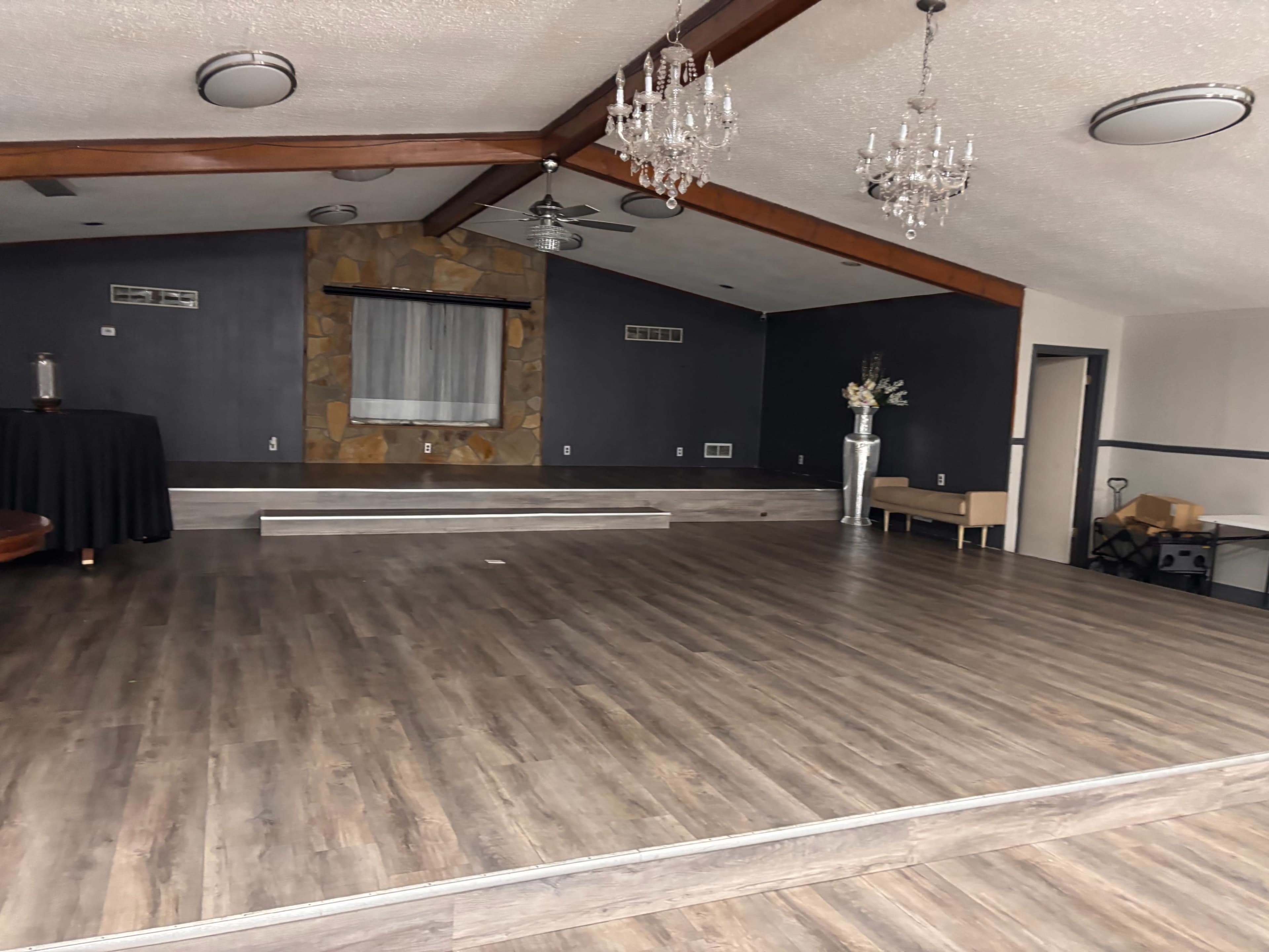 The image shows an empty event space with wooden flooring, a stone accent wall, a stage area, and decorative lighting fixtures hanging from the ceiling.