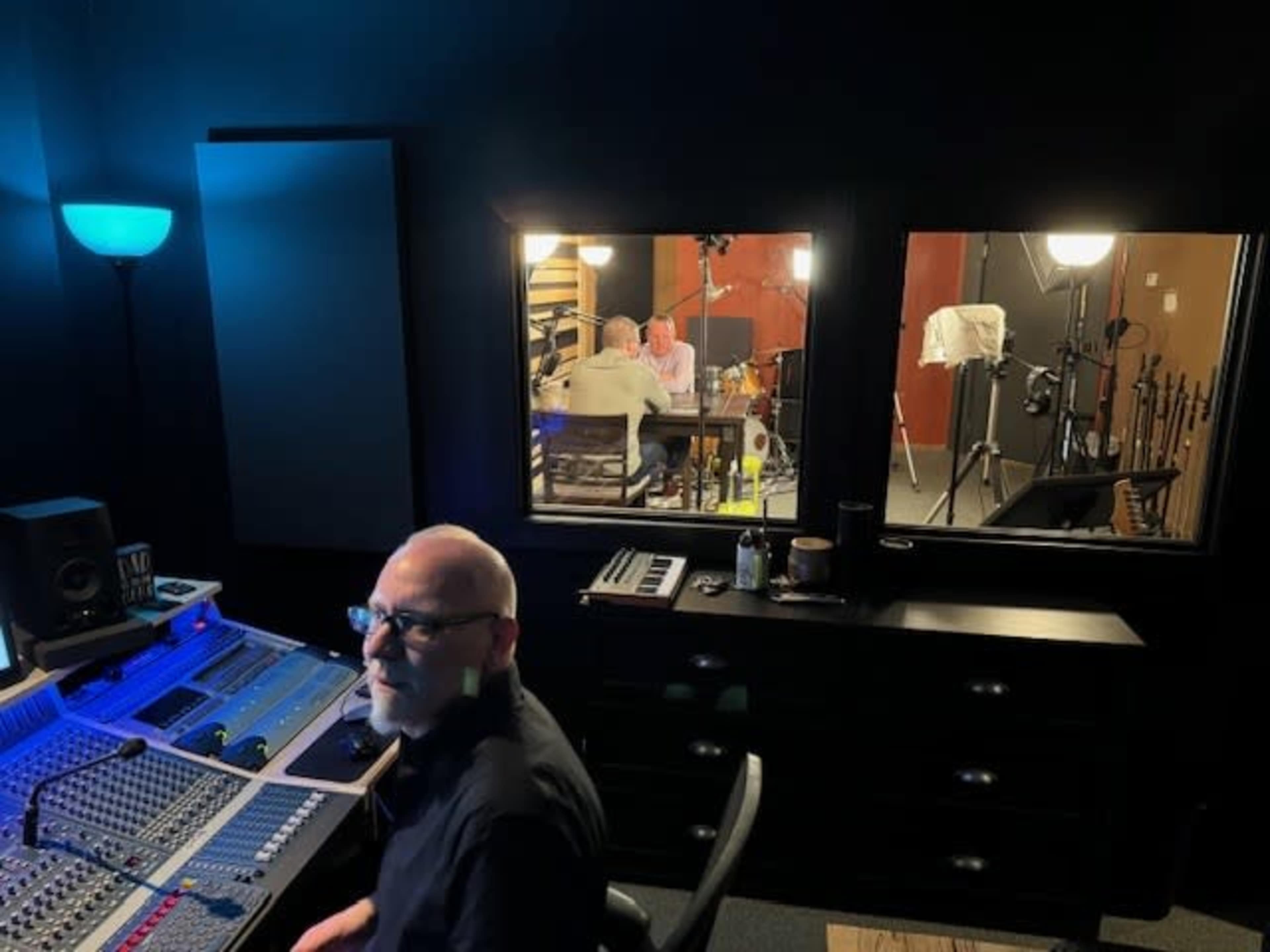 A person sits at a sound mixing console in a dark room, while two individuals are visible through glass windows engaged in a recording session.