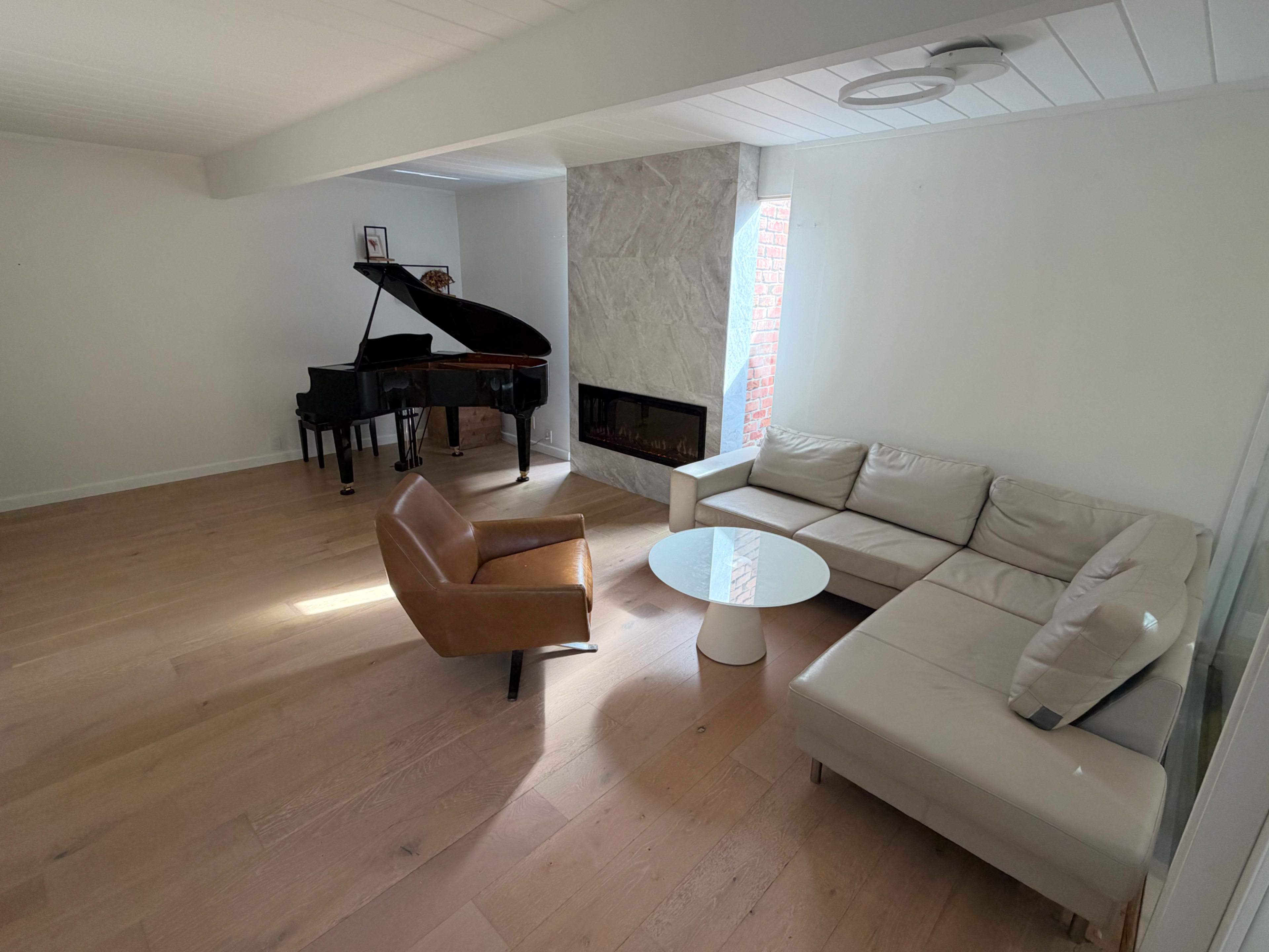 The image shows a modern living room featuring a grand piano, a sectional sofa, a single armchair, and a fireplace.