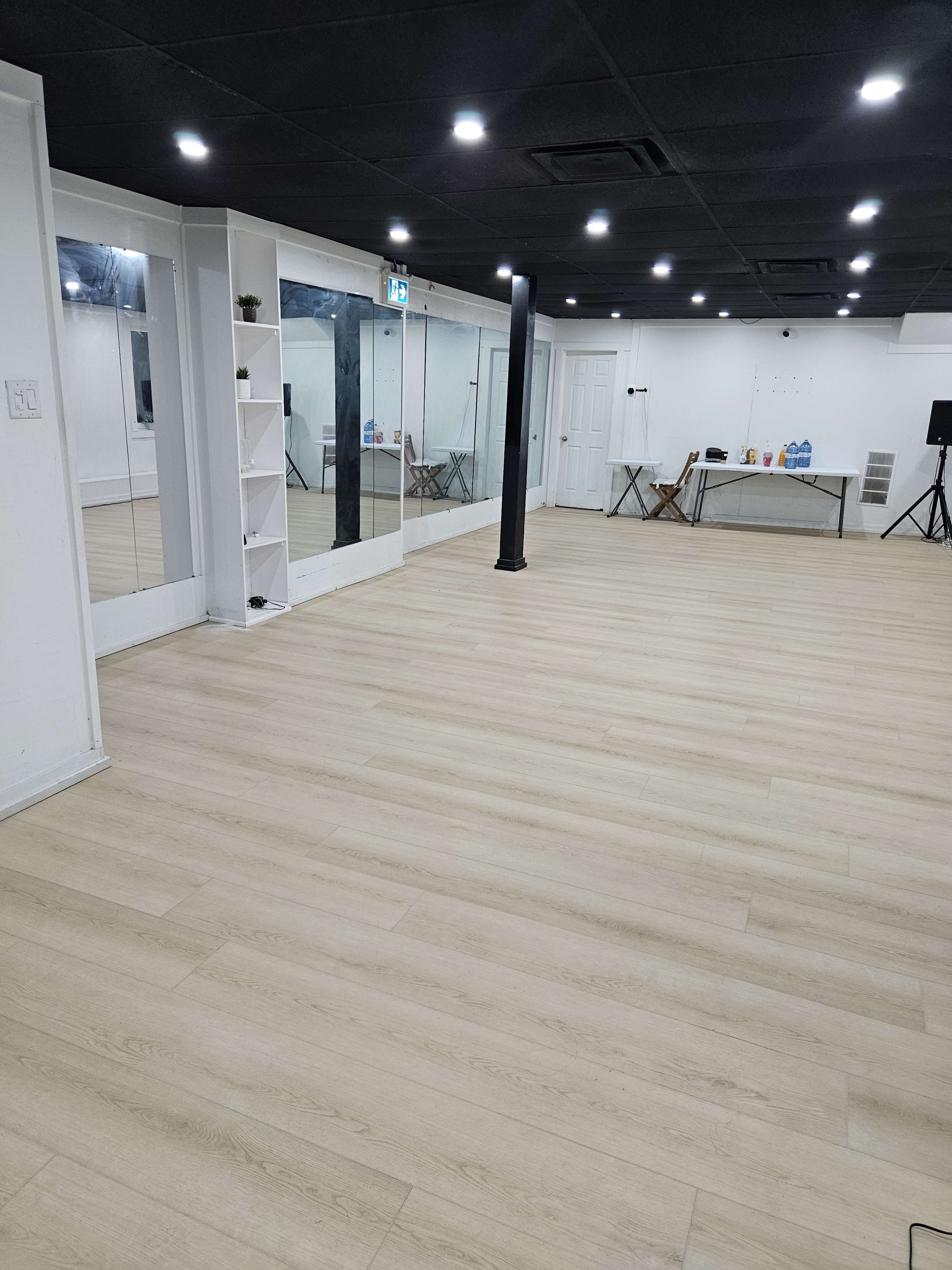 The image shows a spacious, well-lit room with wooden flooring, mirrors on one wall, and minimal furnishings including a table and chairs.