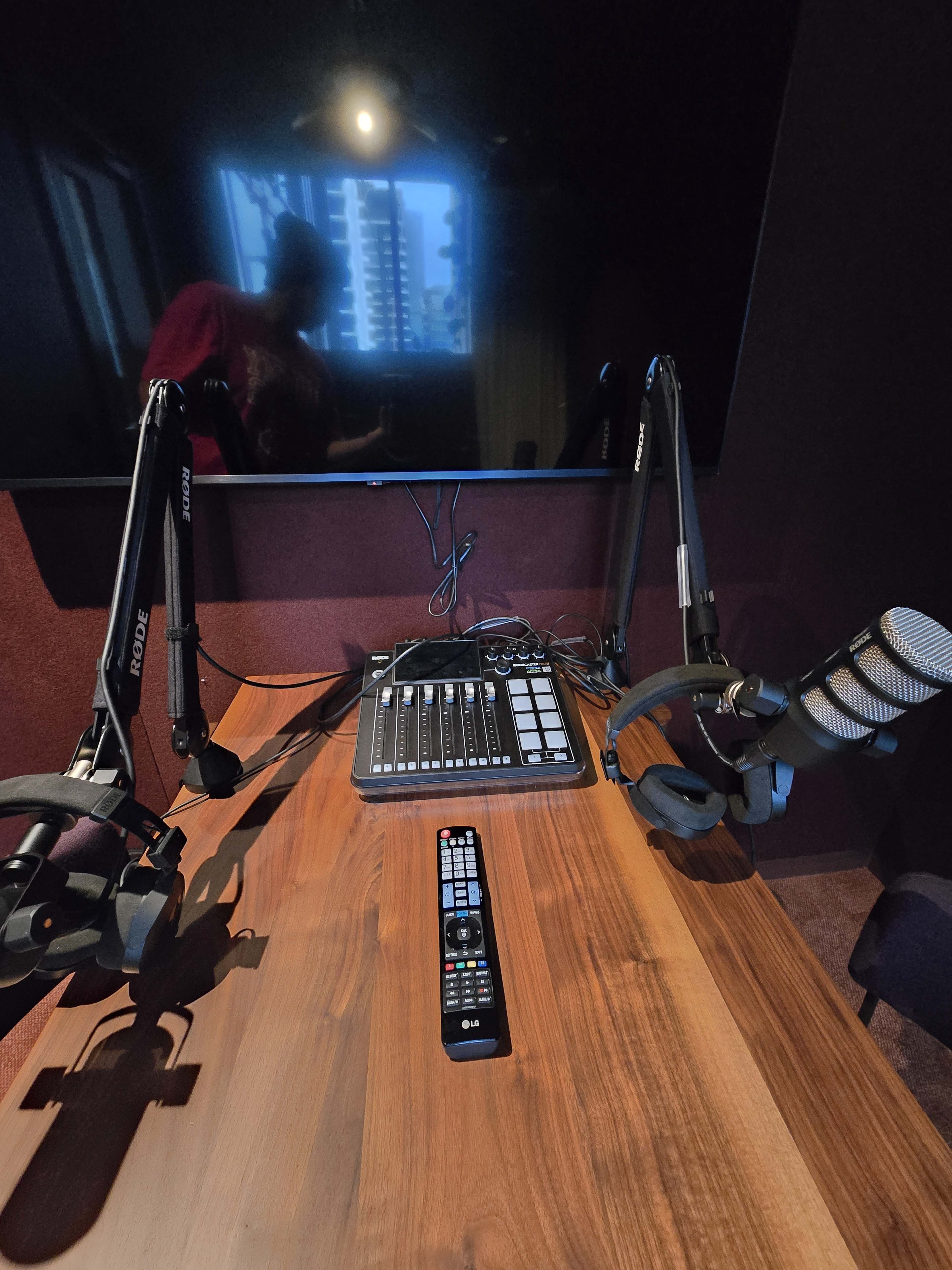 The image shows a radio studio setup with a wooden table, a control panel, two microphone arms, and a remote control on the surface.