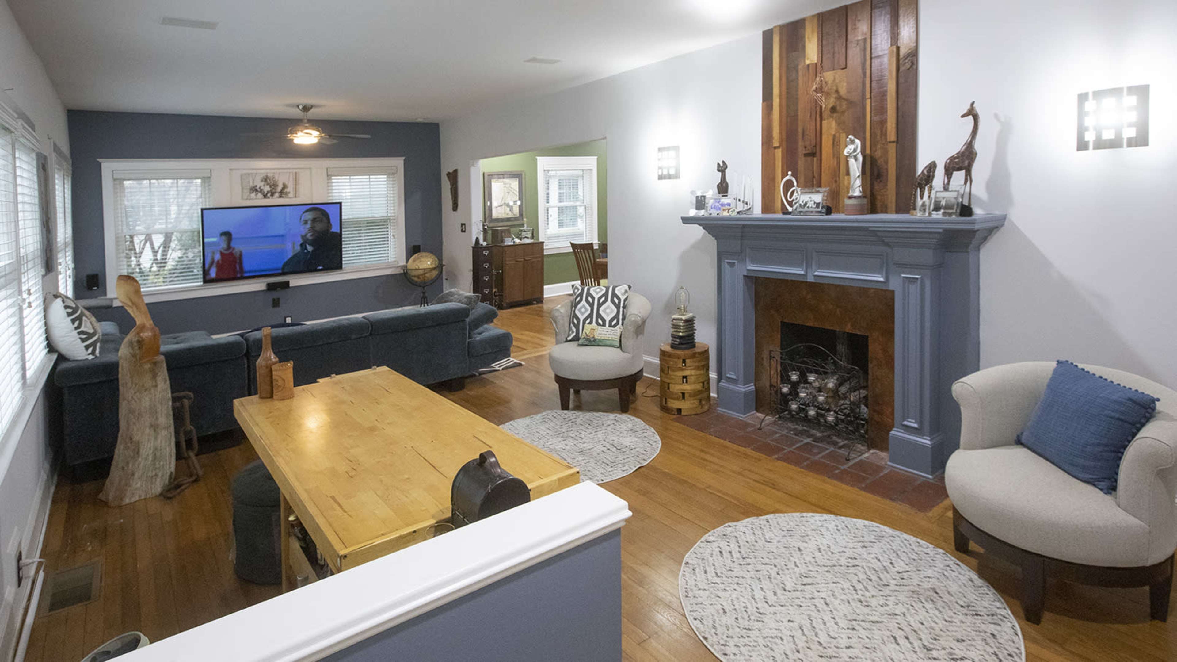 The image shows a living room and dining area with a blue accent wall, a fireplace, a television mounted on the wall, and a wooden dining table surrounded by chairs.