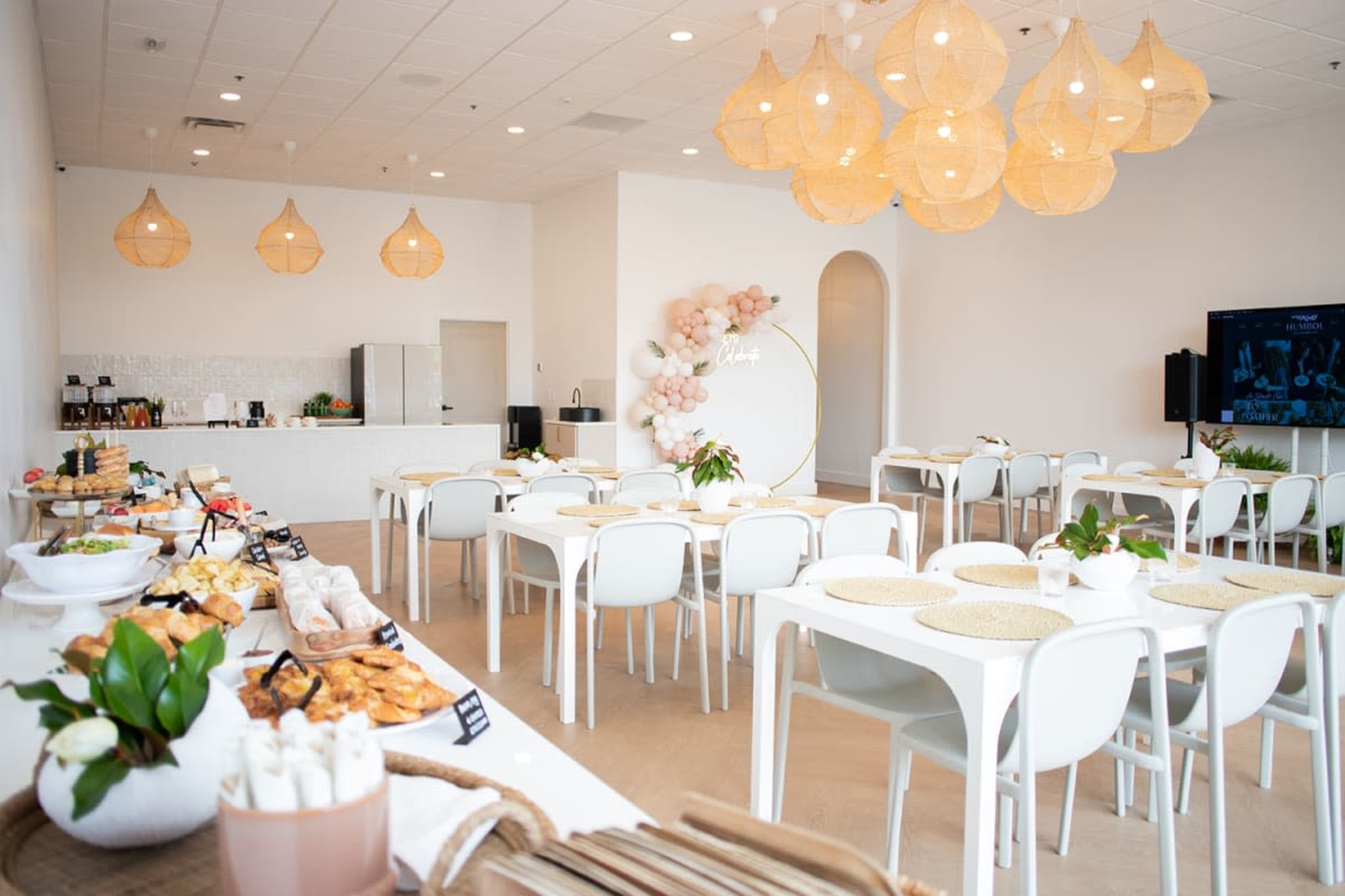 A bright, modern dining area features white tables and chairs, a buffet table with various food items, and decorative hanging lights.