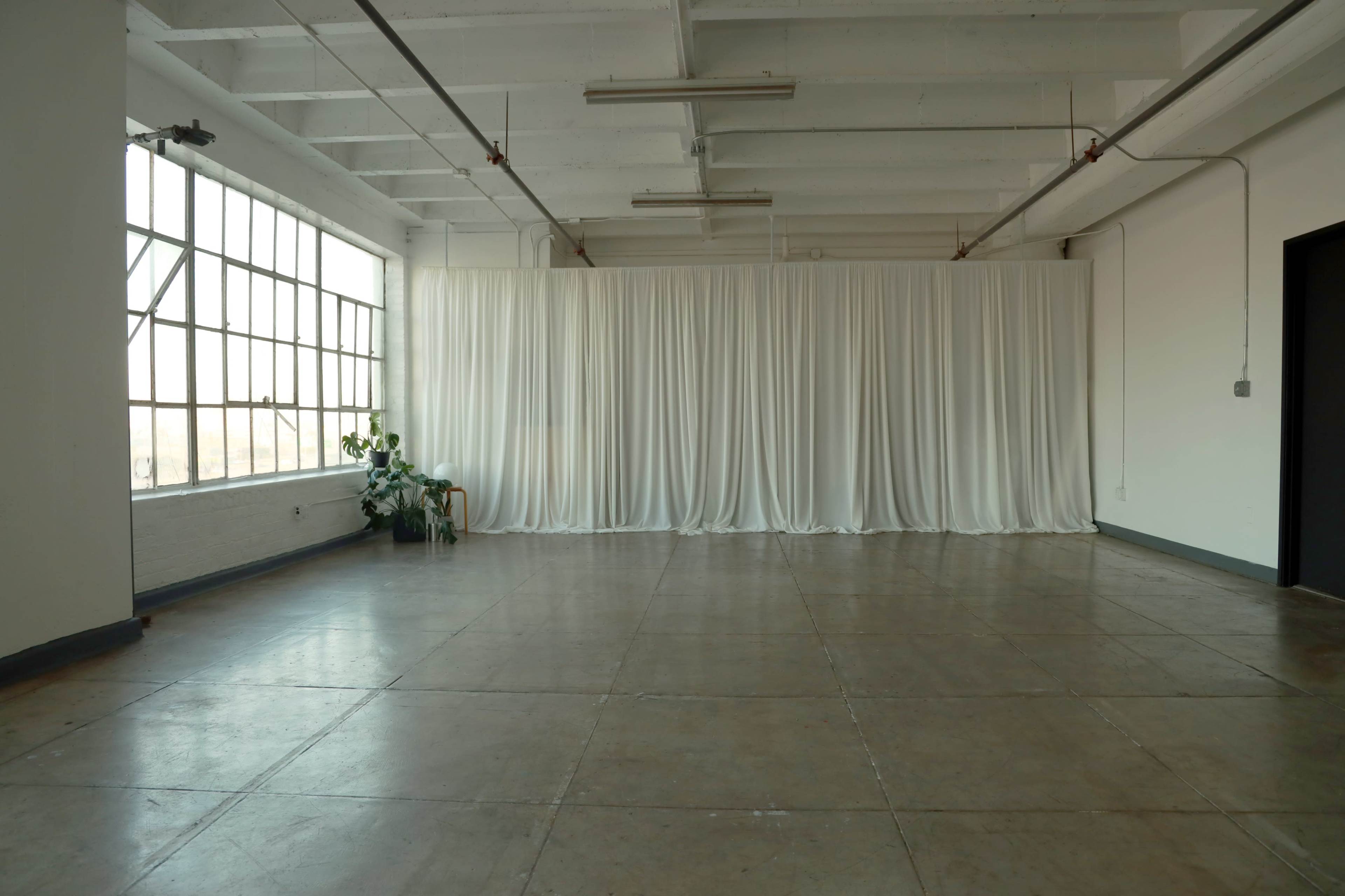 A spacious industrial room features a large window, white drapery along one wall, polished concrete floors, and minimal furnishings.
