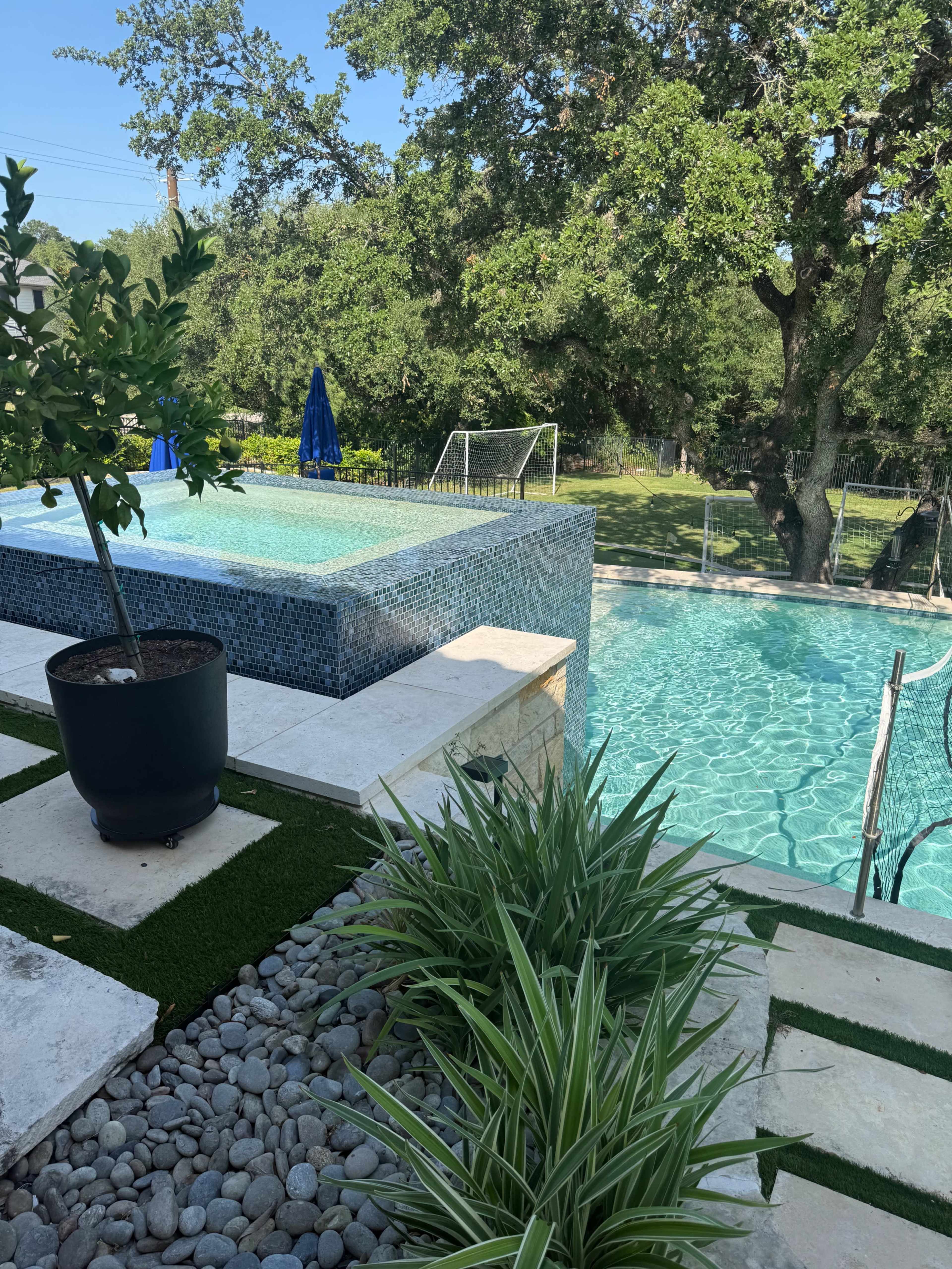 The image shows a landscaped backyard with a swimming pool, a jacuzzi, and green grass under the shade of trees.