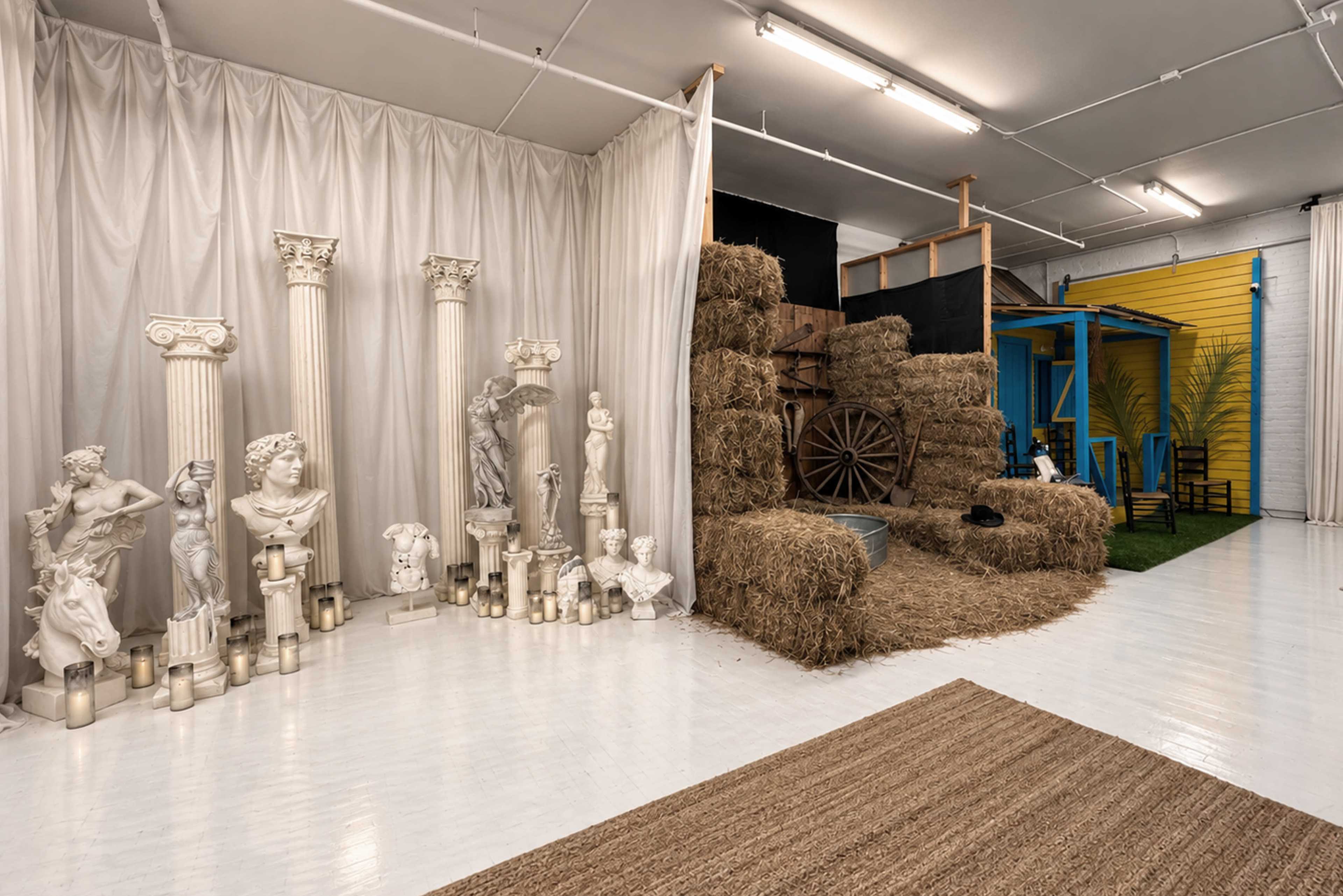 The scene features a studio space with decorative classical sculptures and columns on one side, adjacent to a rustic area made of hay bales and a colorful structure resembling a house.
