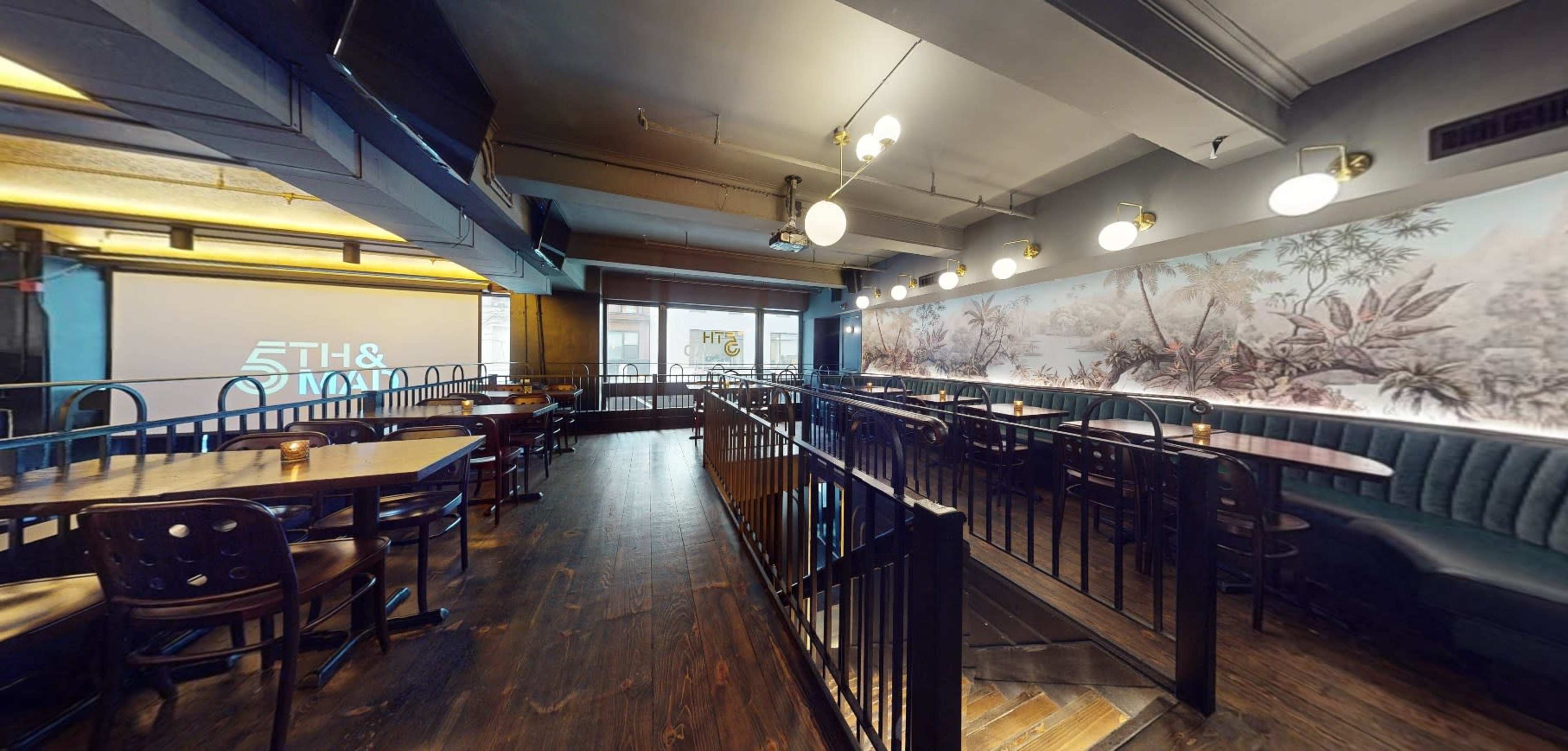 The image shows a spacious restaurant interior with wooden tables and chairs, featuring a large mural on one wall and a projector screen on another.