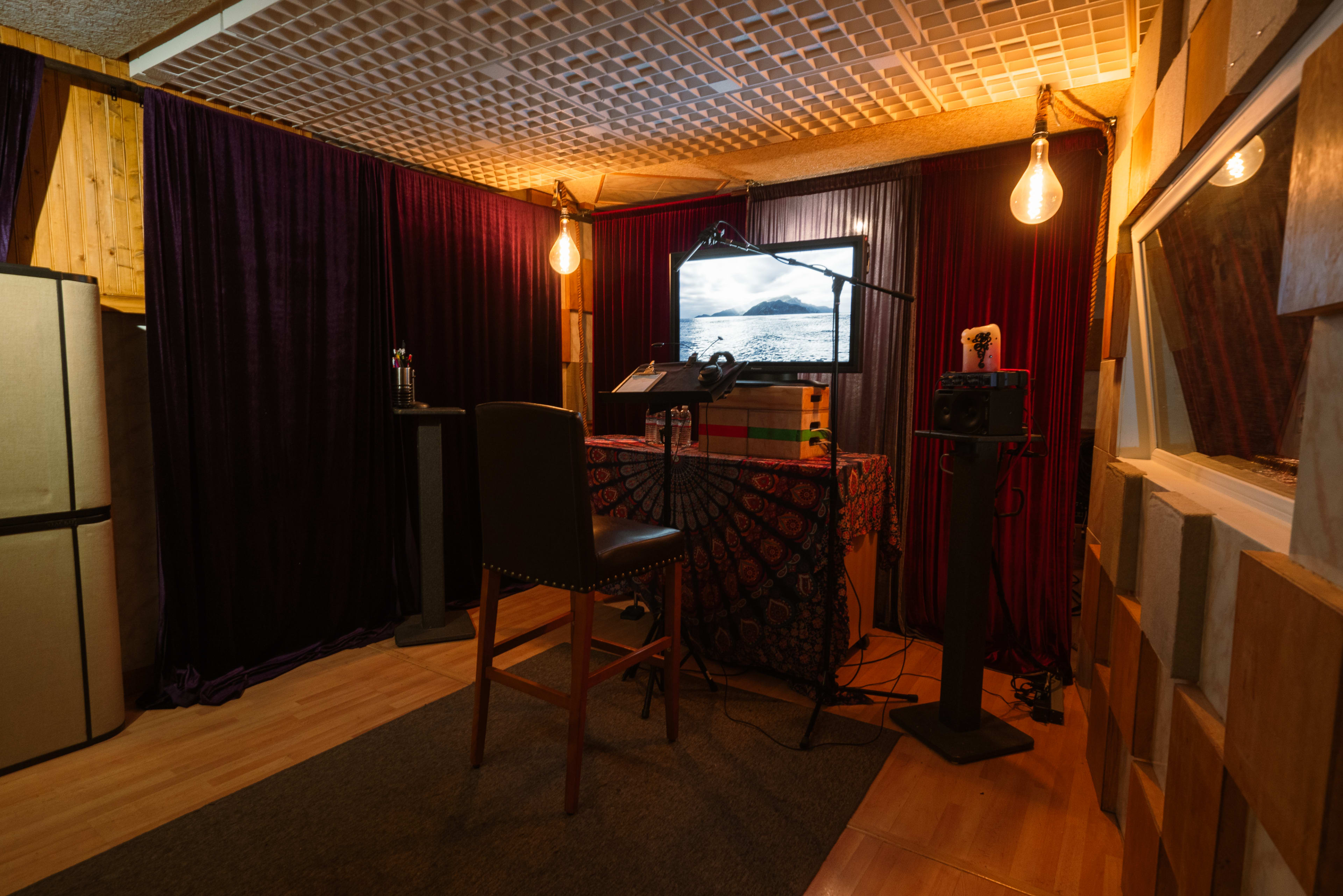 The image shows a dimly lit recording studio featuring a desk with a monitor, microphone, and audio equipment, surrounded by draped fabric and acoustic panels.