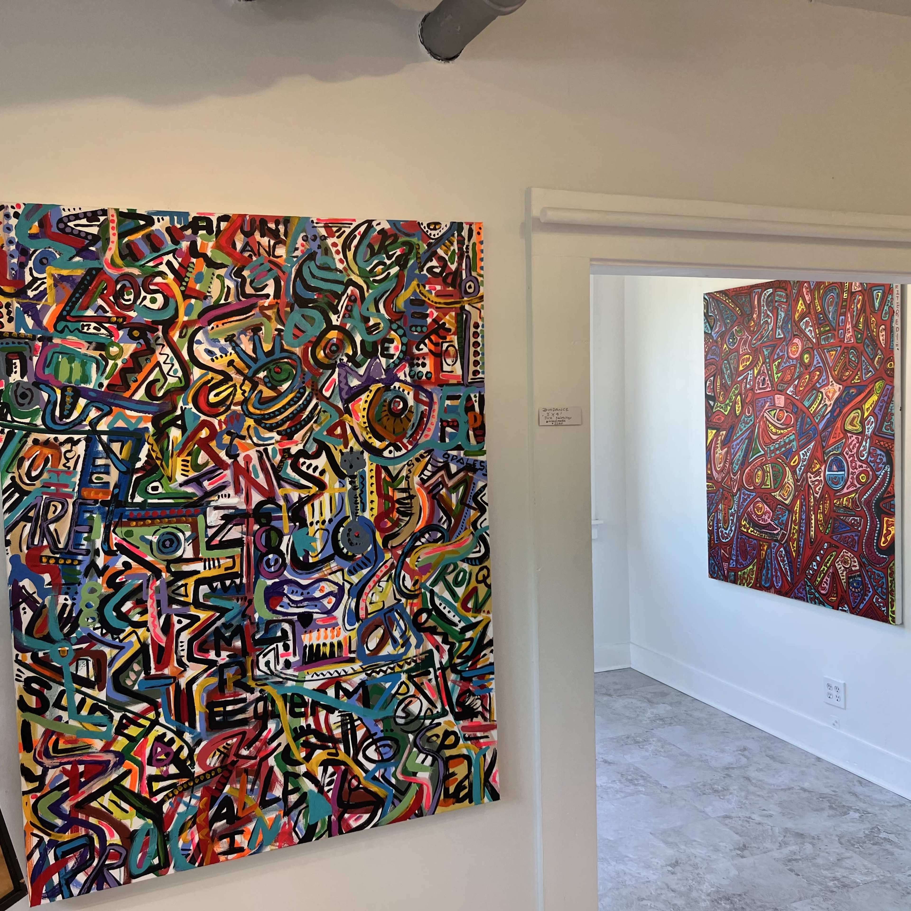 The image shows two abstract paintings displayed on the walls of a bright room with light-colored flooring.
