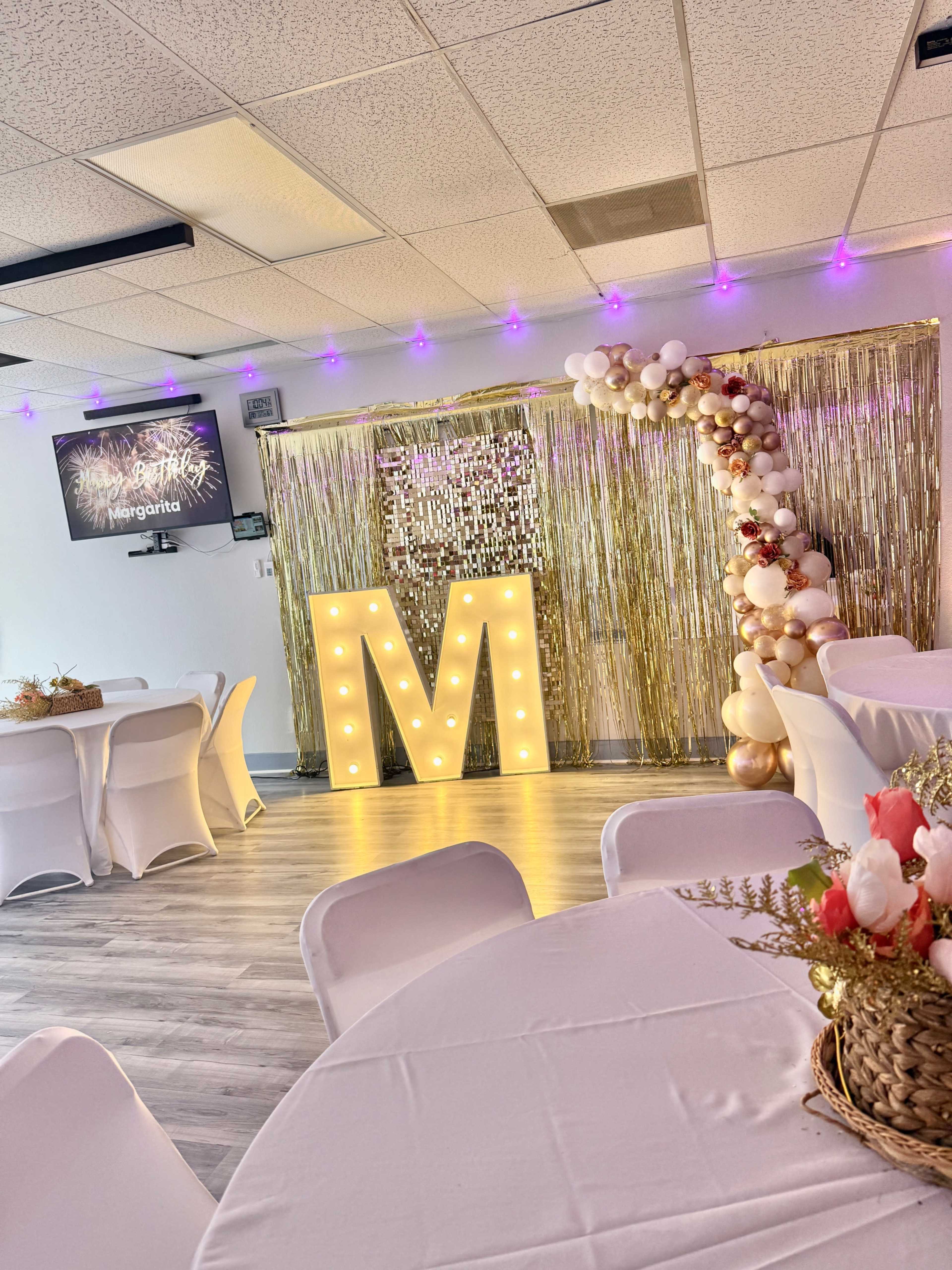 The image shows a decorated event space featuring a large illuminated letter "M," a backdrop with gold fringe, and balloon arrangements.