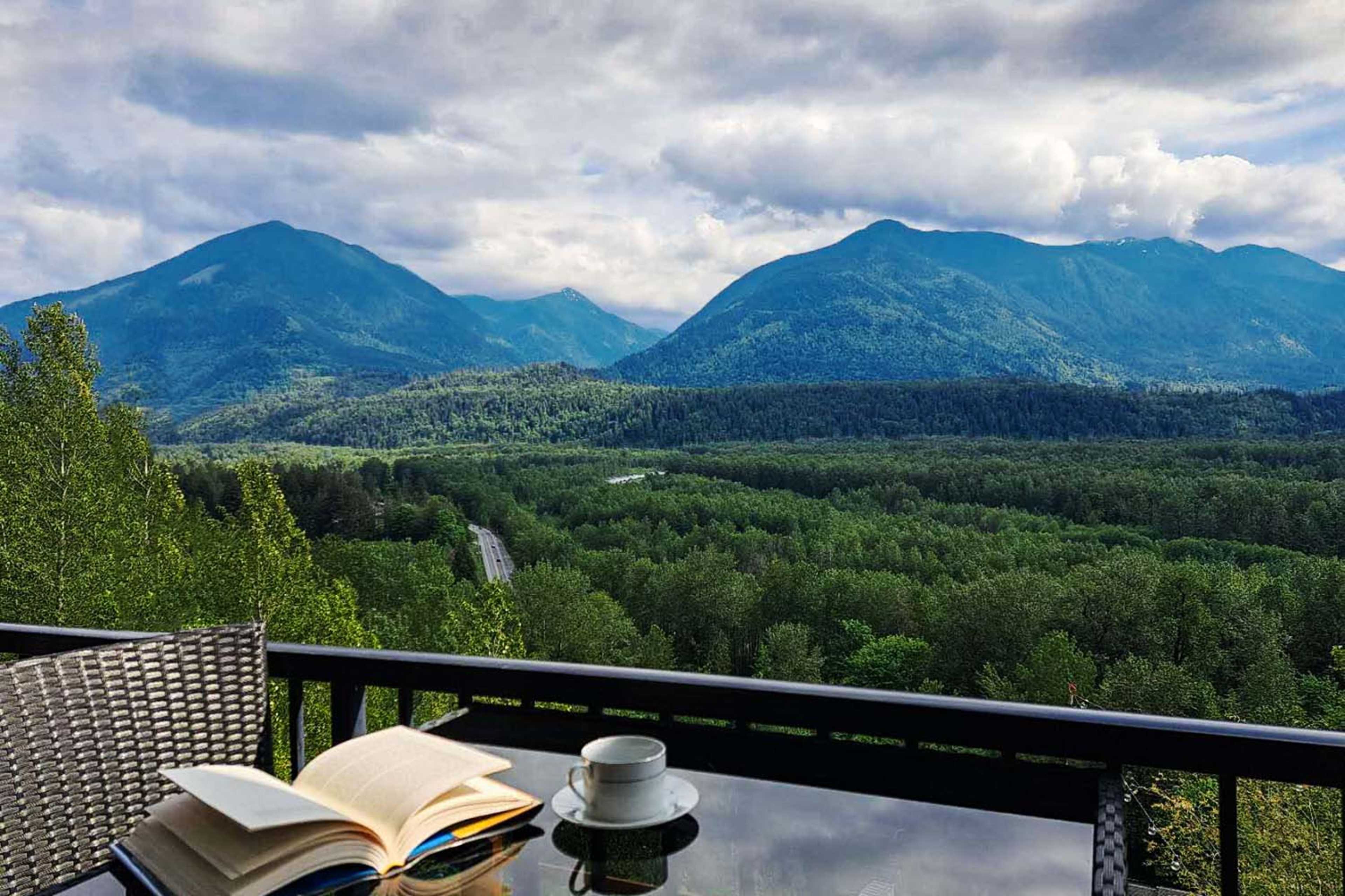 An open book and a coffee cup sit on a table overlooking a lush green valley and distant mountains under a cloudy sky.