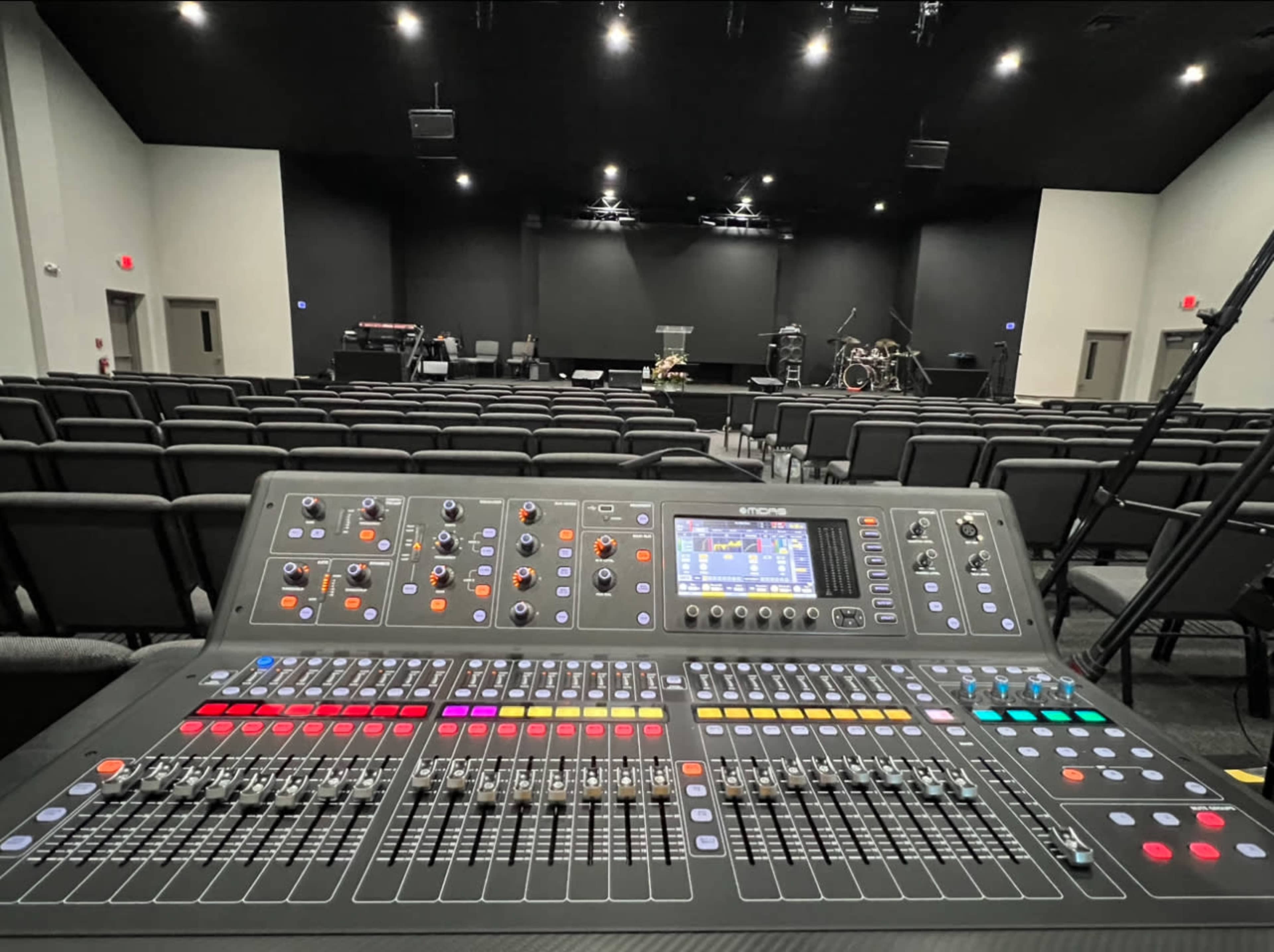 A digital sound mixing console is in the foreground with an empty auditorium and stage setup visible in the background.