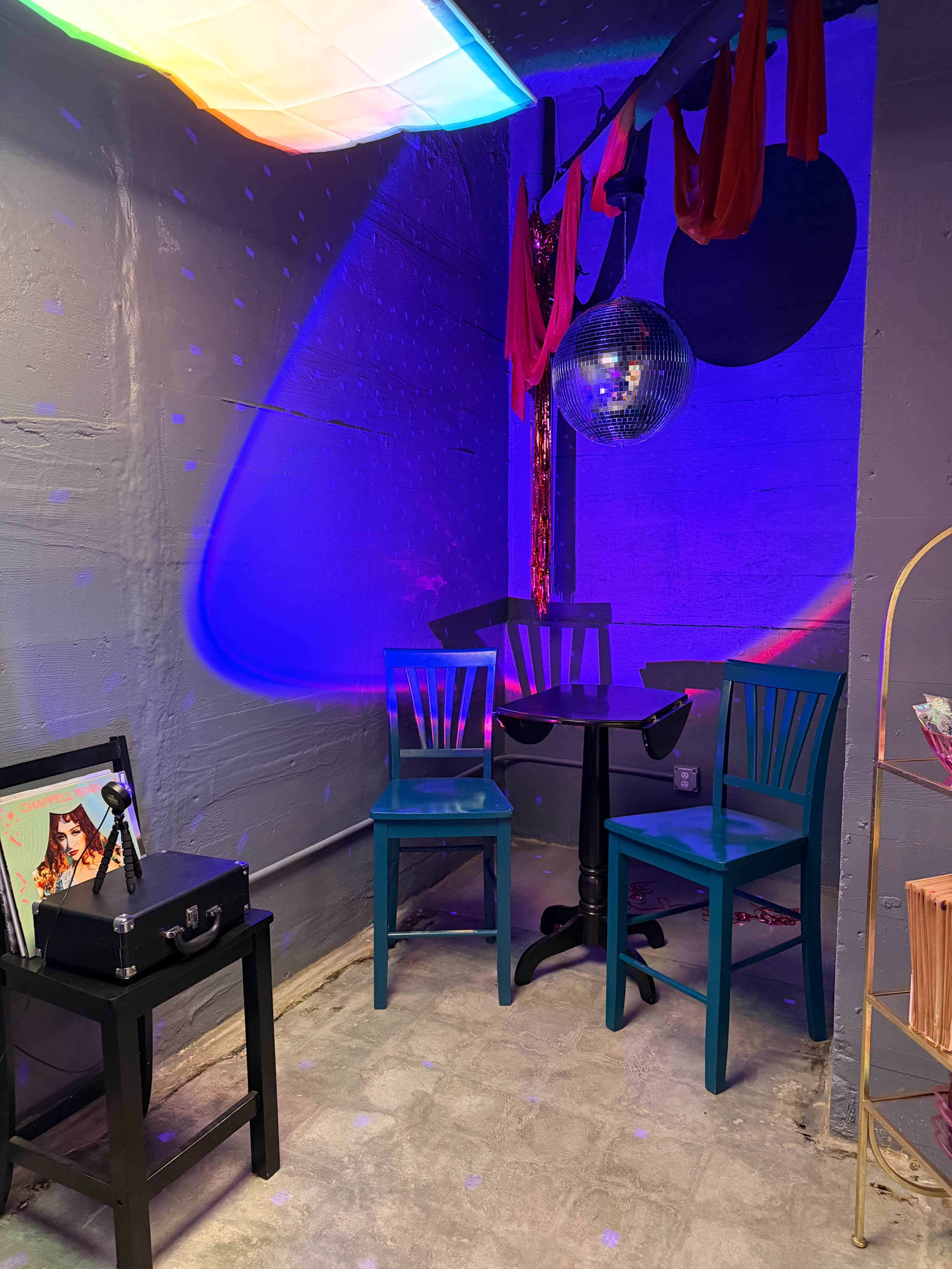 The image shows a small room featuring a disco ball, colorful lighting, two teal chairs, and a black table, creating a vibrant atmosphere.