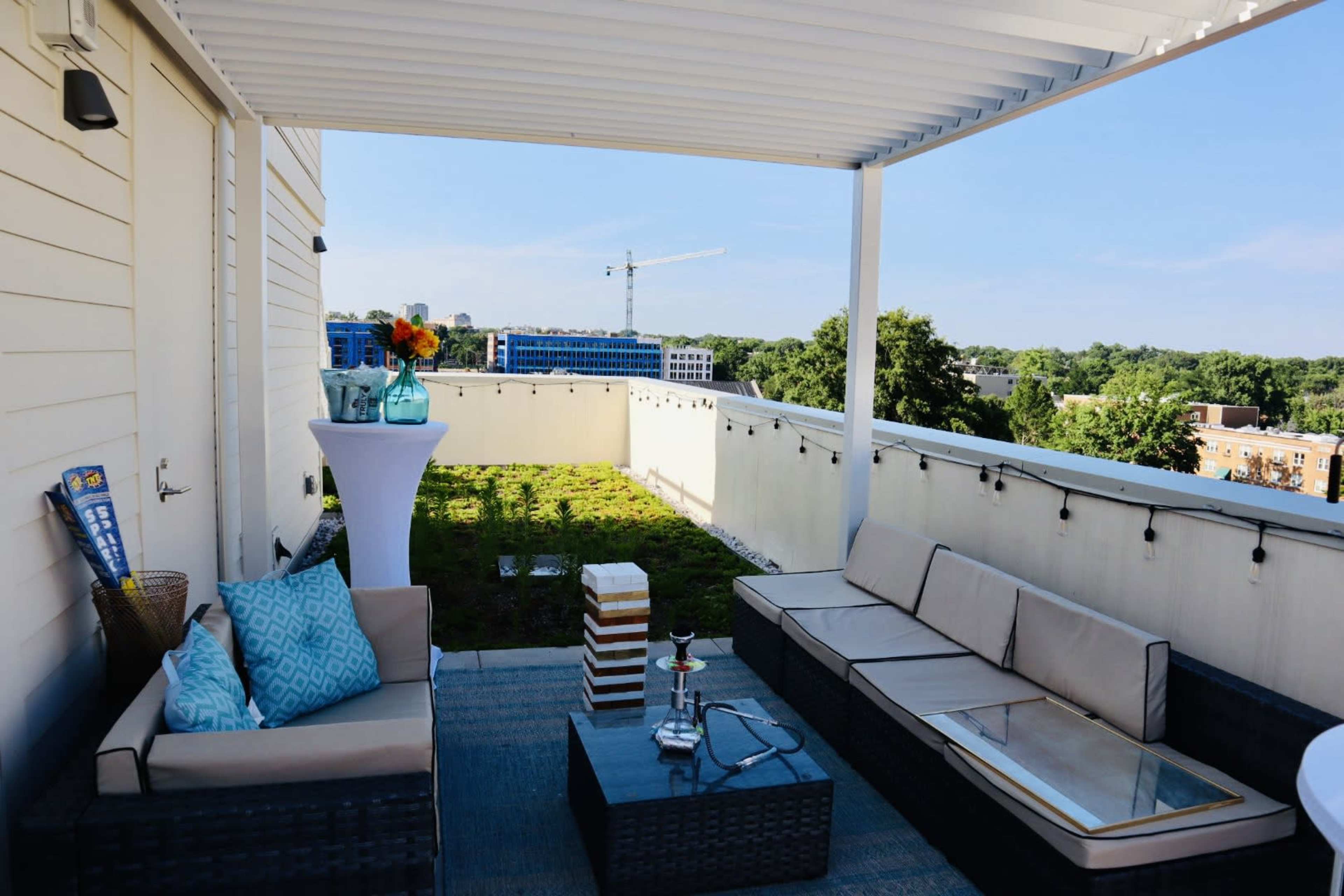 The image shows a rooftop terrace with modern seating, a table, and a small garden area, overlooking a city landscape.