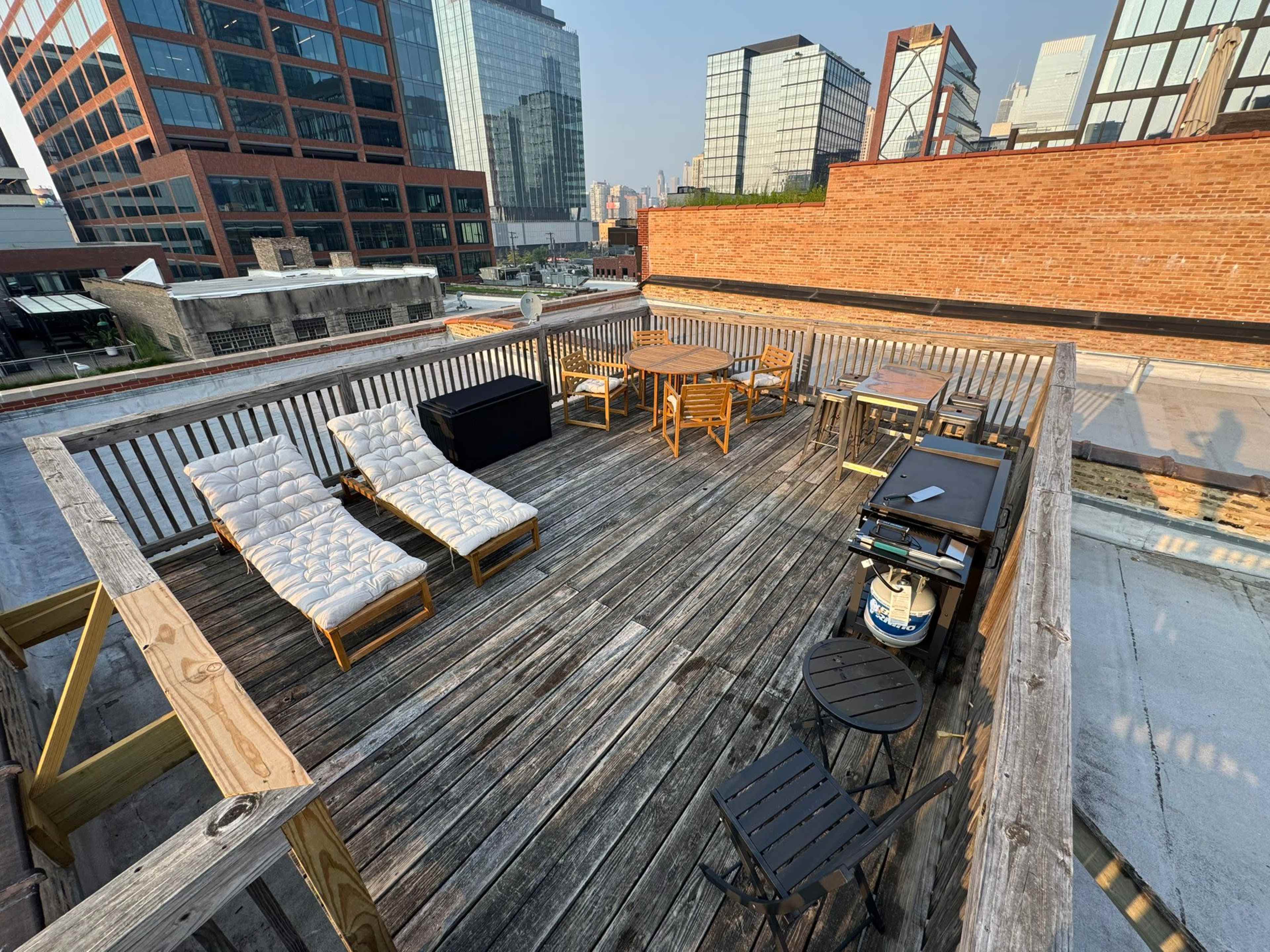 The image shows a rooftop deck with wooden flooring featuring several pieces of outdoor furniture, including lounge chairs, a table, and a grill, surrounded by tall buildings.