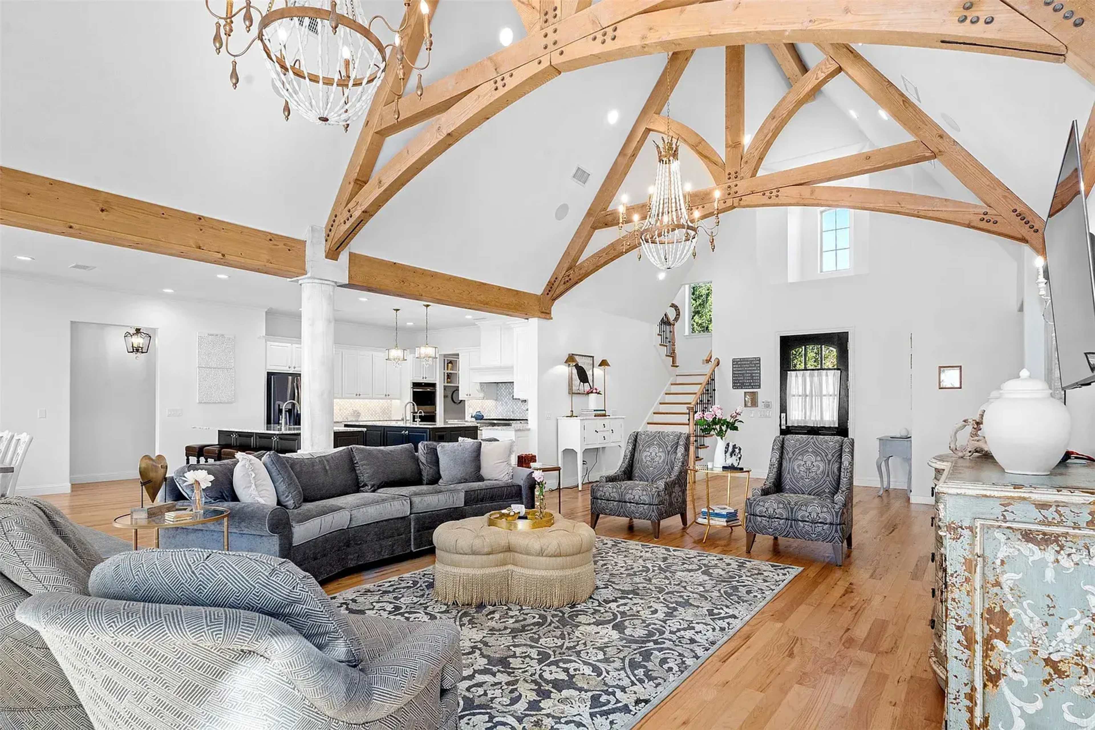 A spacious living room features a high, wooden beam ceiling, large windows, and a mix of modern and traditional furnishings.