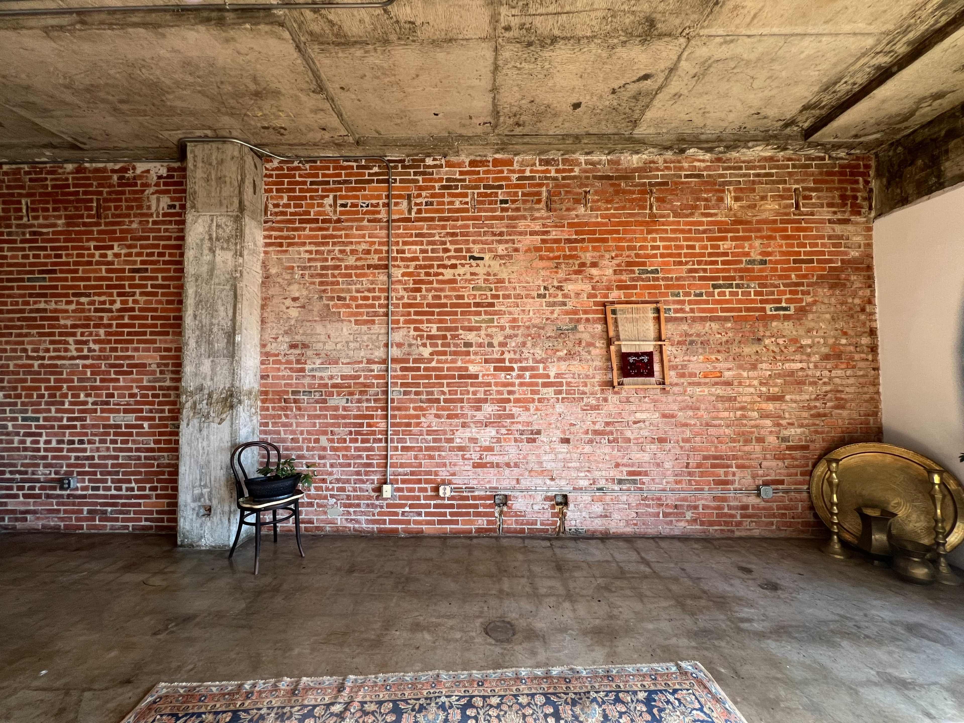 The image shows an industrial-style room with a brick wall, a wooden chair with a plant next to it, and a decorative rug on a concrete floor.