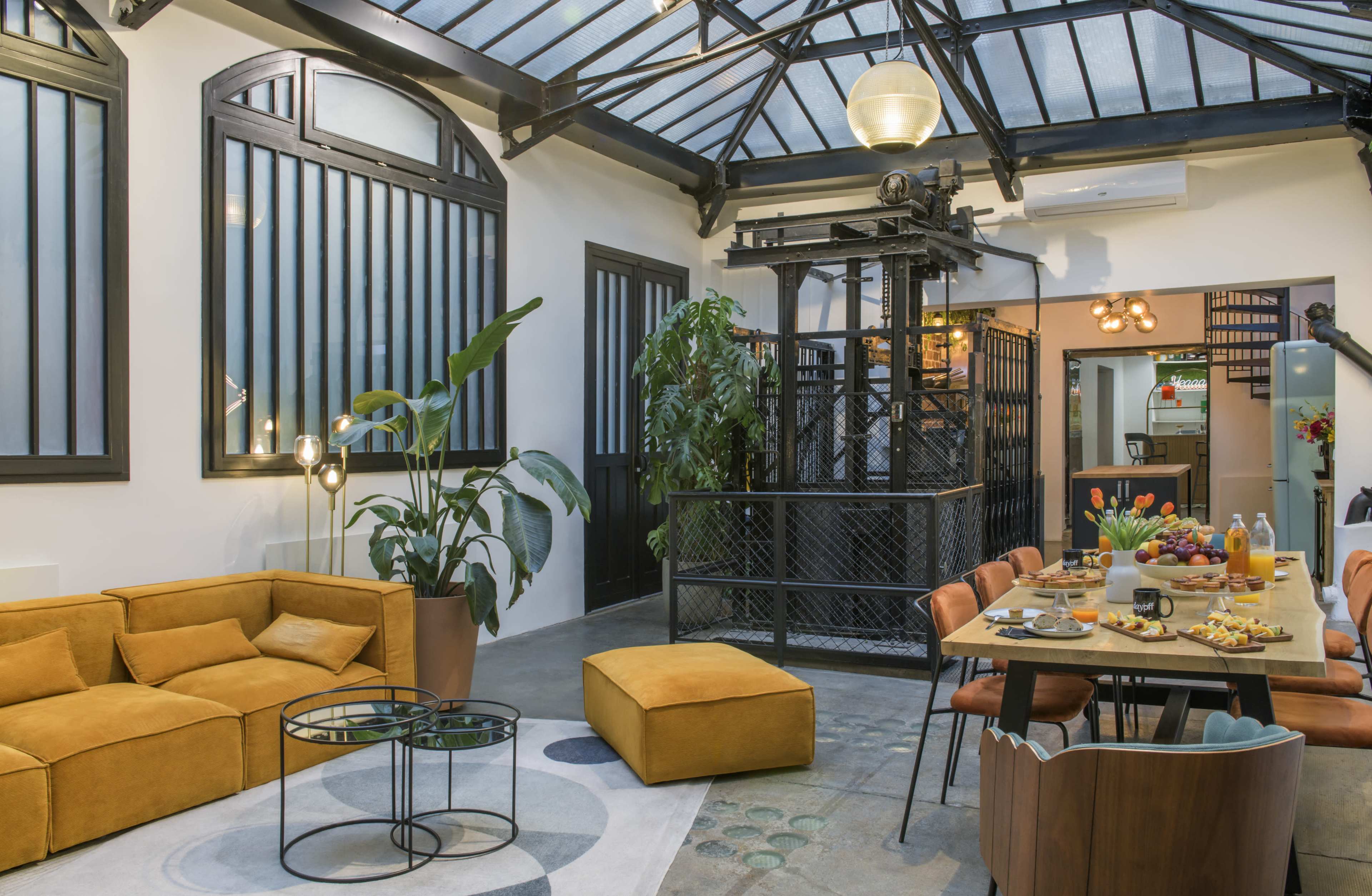 The image shows a modern, open-concept living space with a kitchen area, featuring a mix of stylish furniture, plants, and decorative elements, alongside an antique lift.