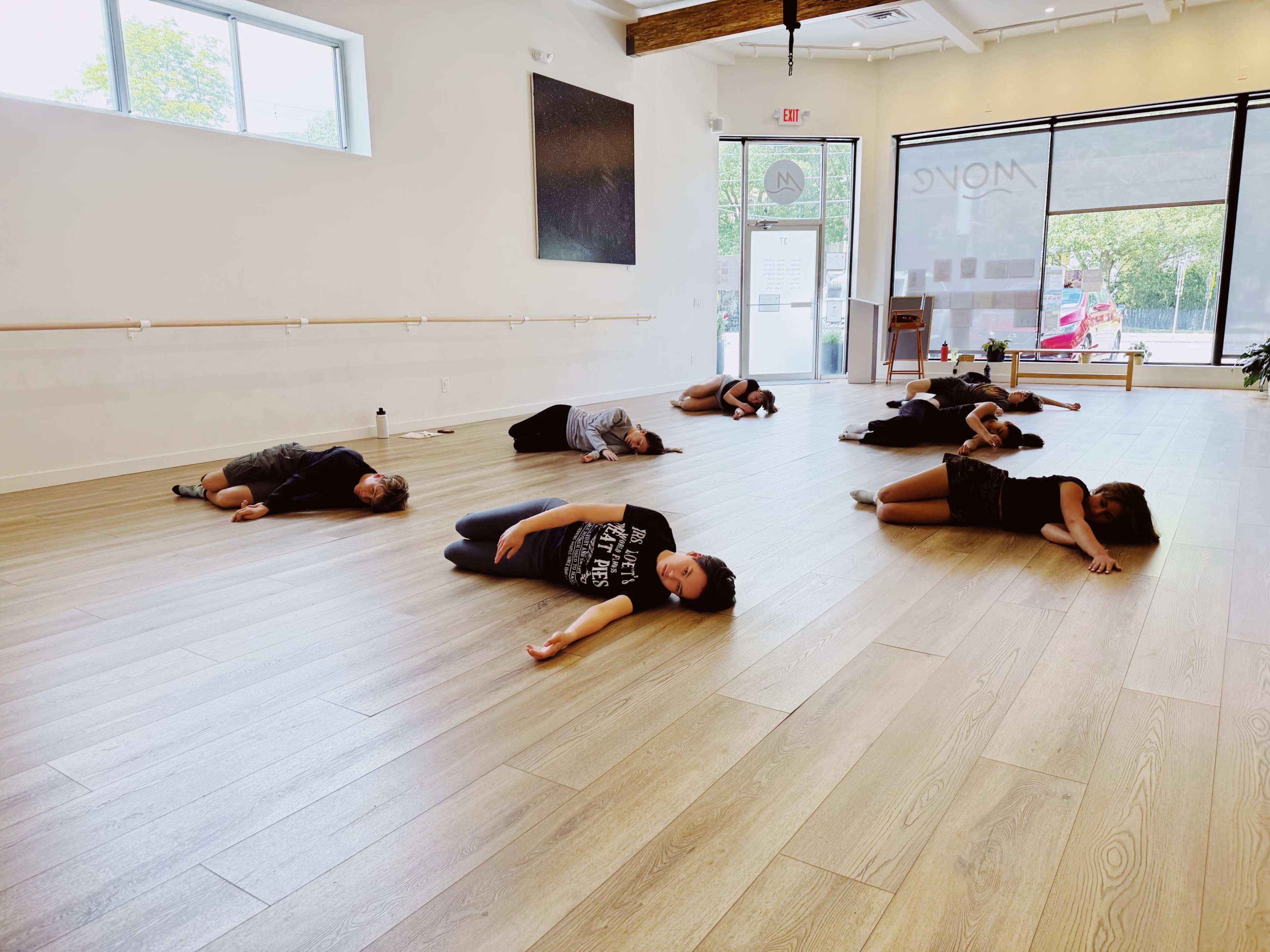 A group of people is lying on the wooden floor of a spacious dance studio.
