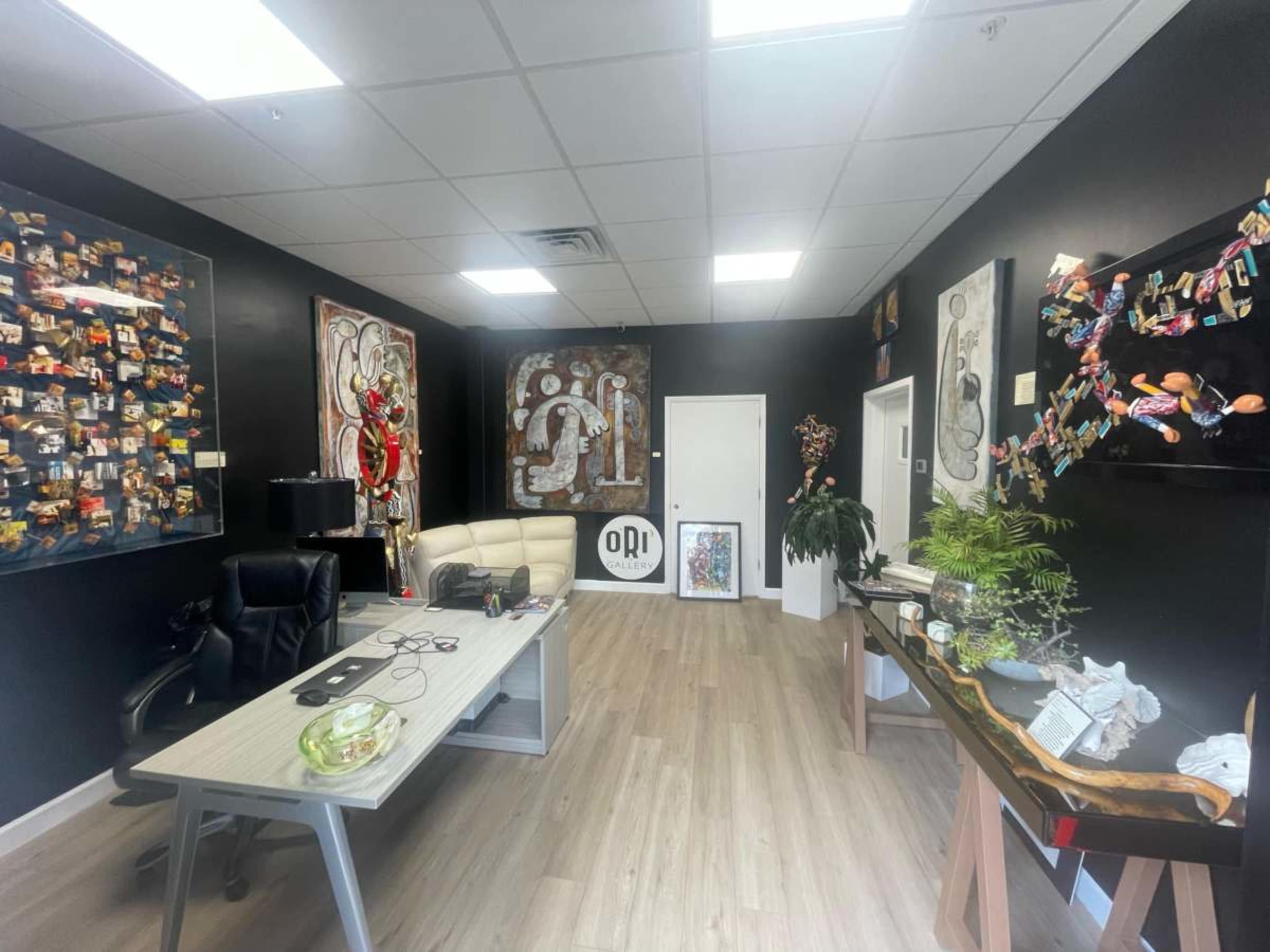 The image shows a modern office space featuring a black wall adorned with colorful artwork, a light-colored desk with a computer chair, and various decorative elements, including plants and a gallery of framed photographs.