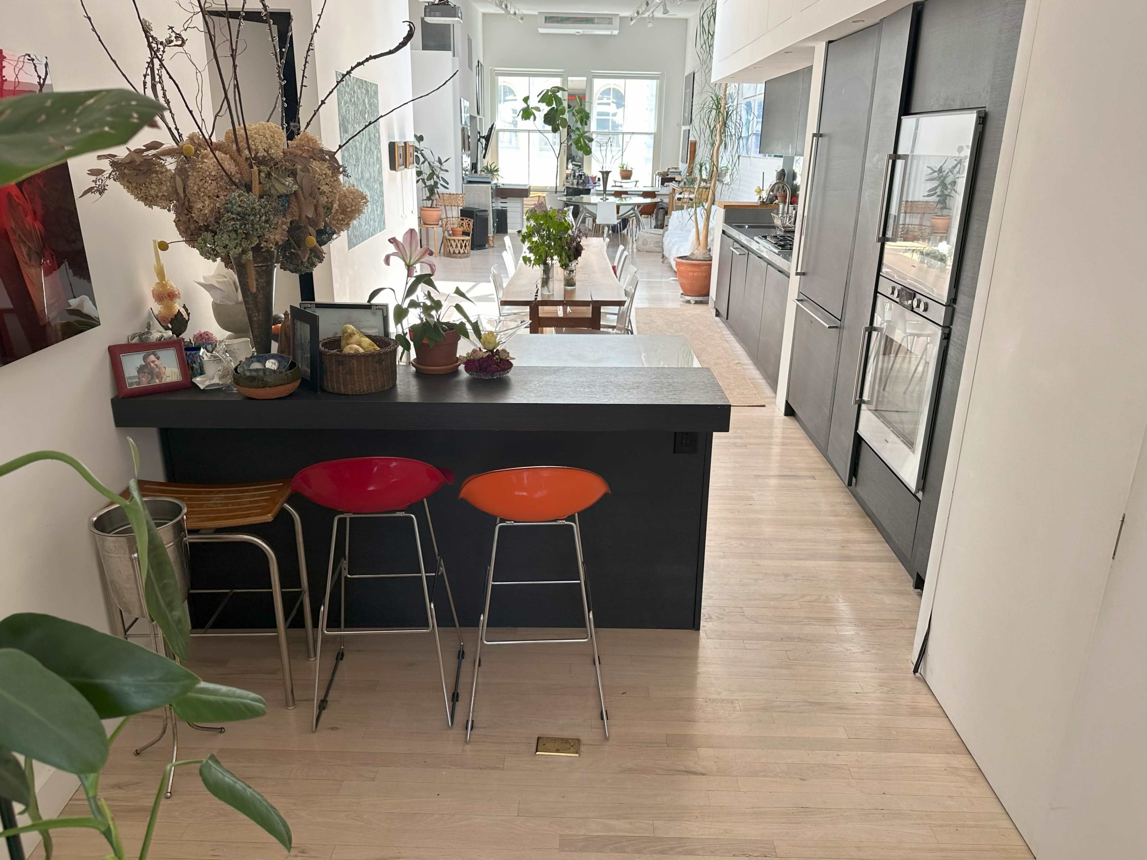 A modern kitchen with a minimalist bar area featuring three stools, surrounded by plants and a dining area in the background.