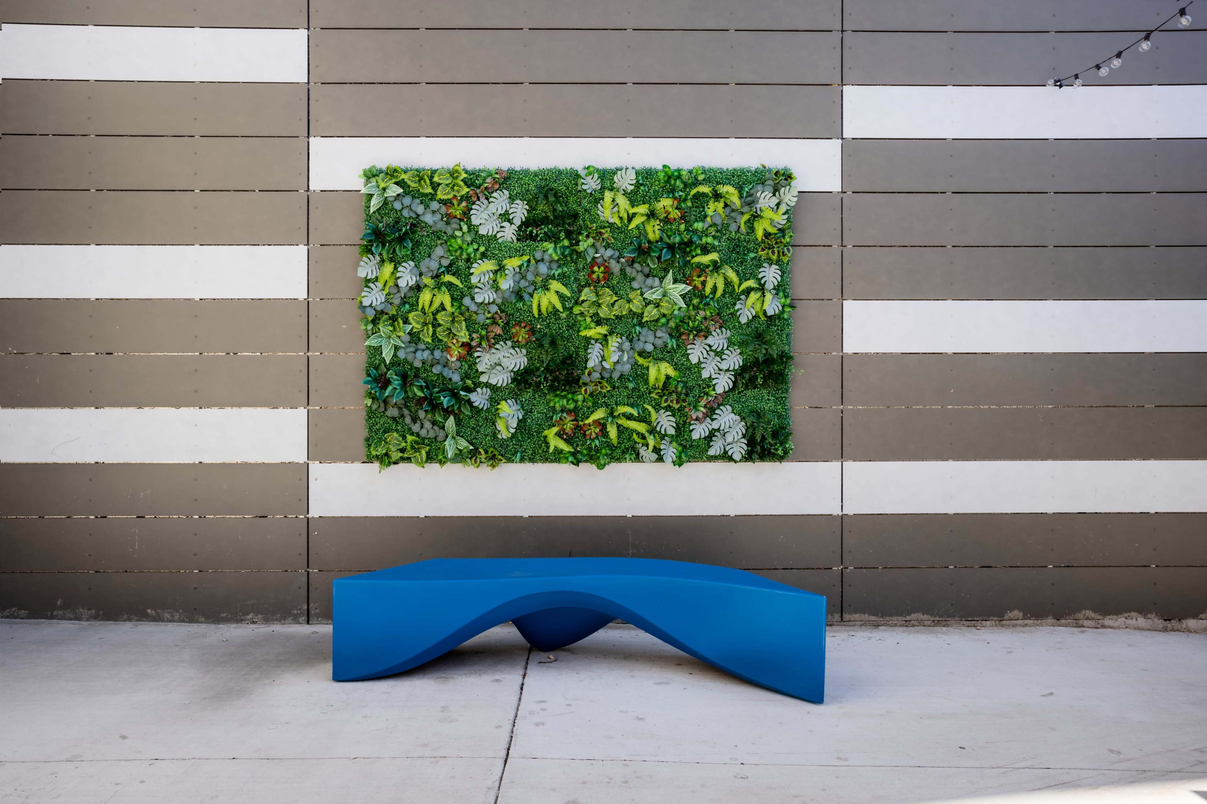 A blue sculptural bench is placed in front of a vertical garden on a gray and white striped wall.