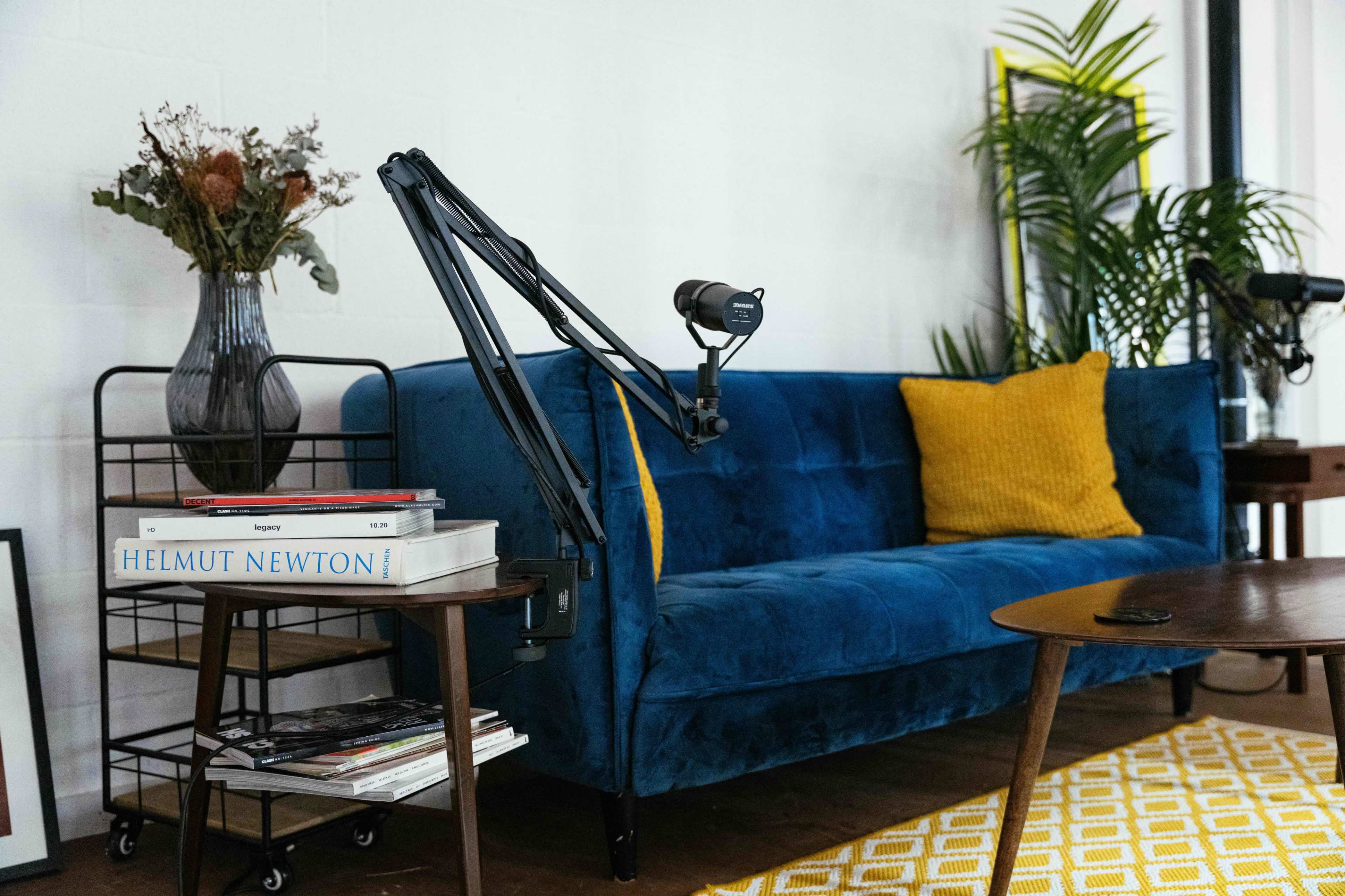 A blue sofa with a yellow pillow, a side table stacked with books, and a microphone on an arm in a modern interior setting.