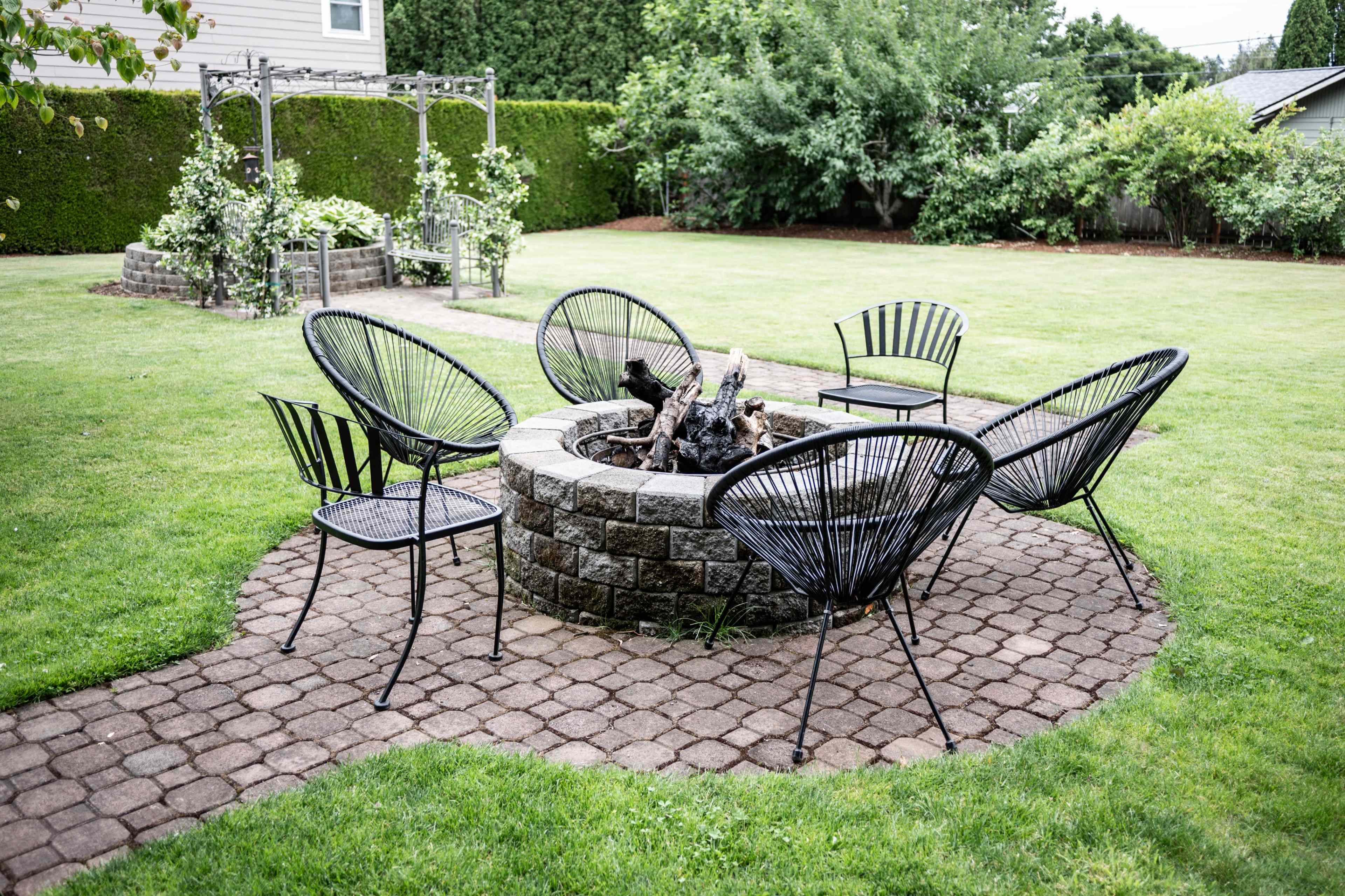 A circular stone fire pit is surrounded by several black chairs in a grassy backyard.