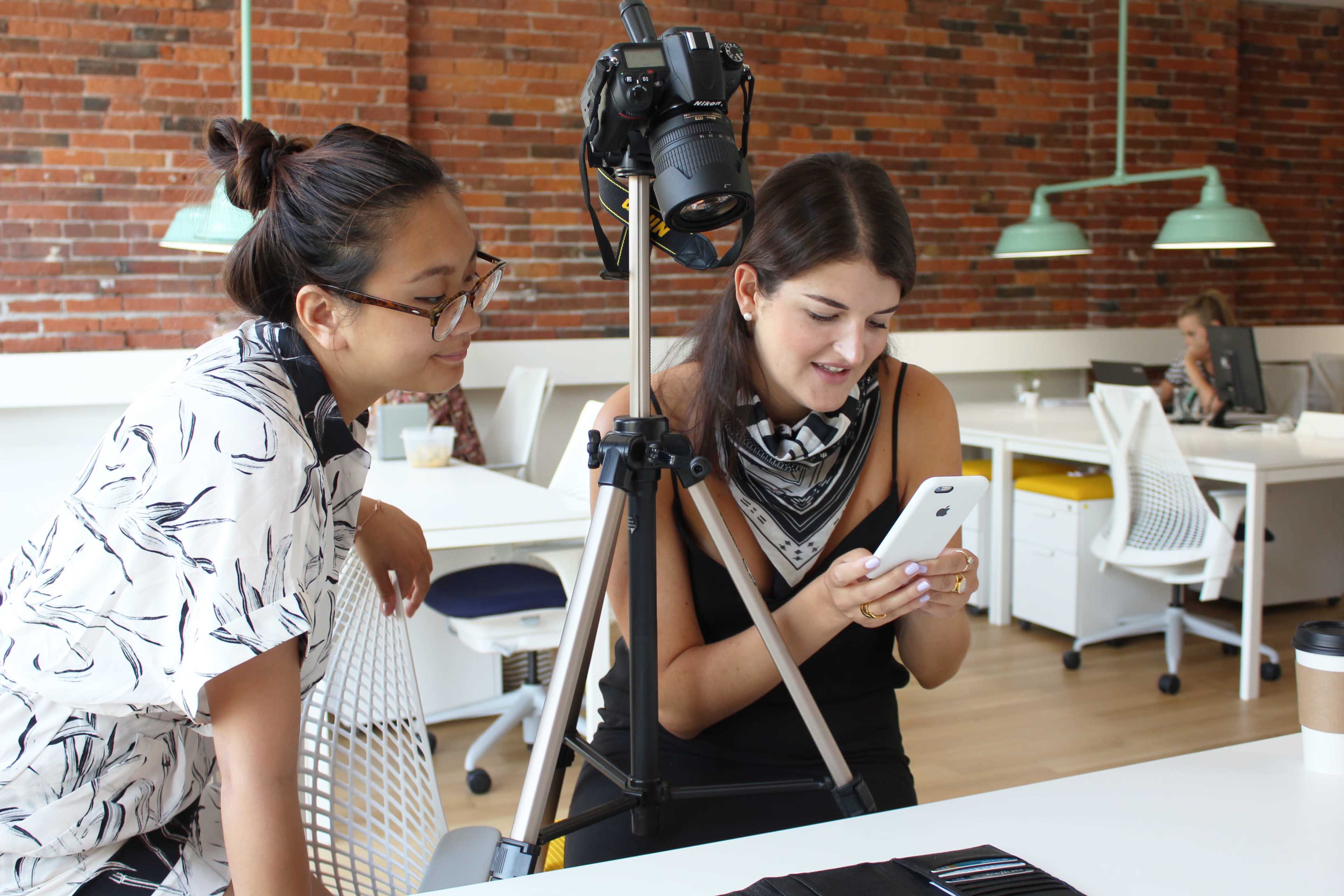 Two women are in an office with a camera mounted on a tripod, one checking her phone while the other looks on.