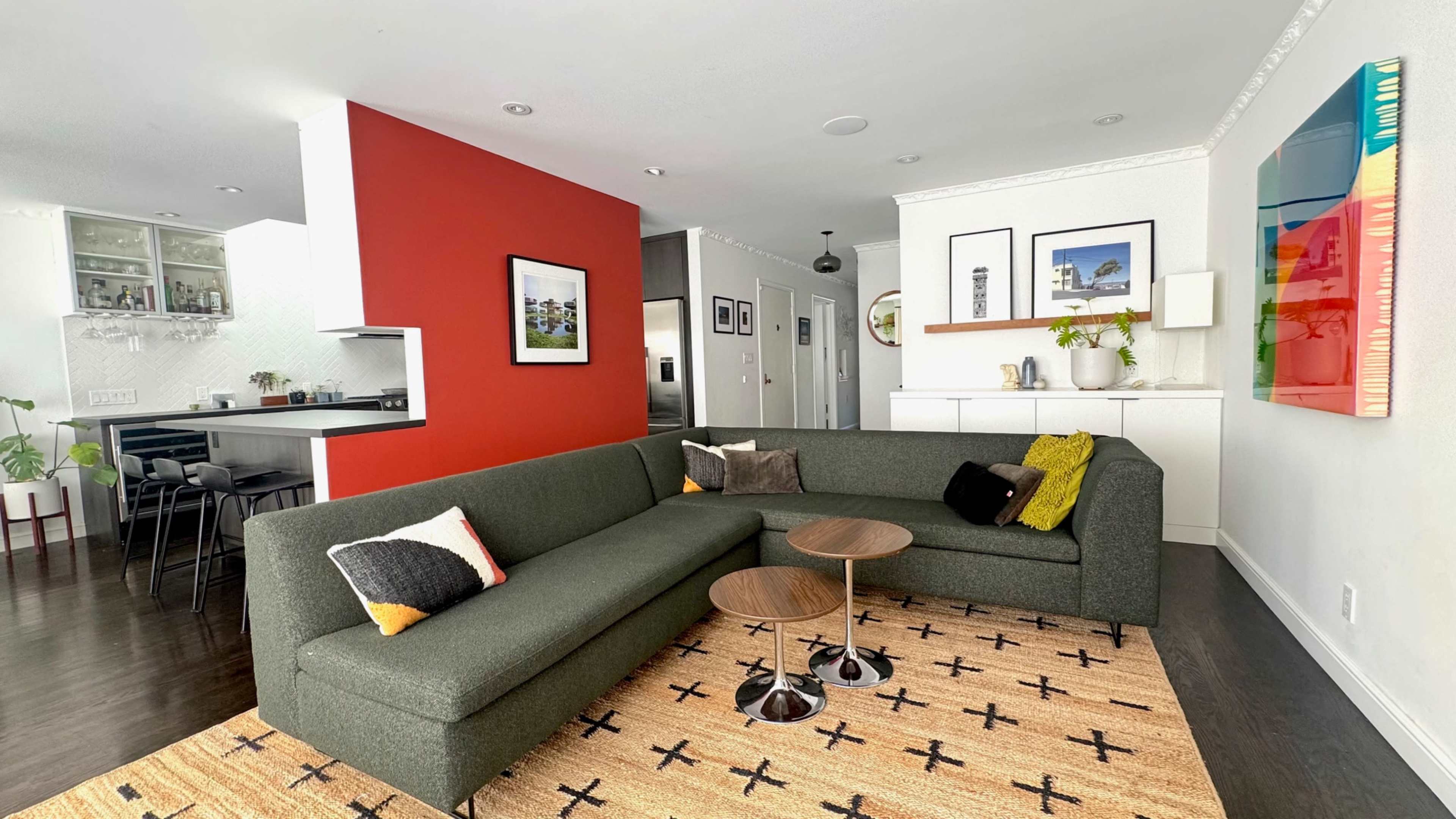 The image shows a modern living room with a dark green sectional sofa, two small tables, and a patterned rug, complemented by colorful wall art and an open kitchen in the background.