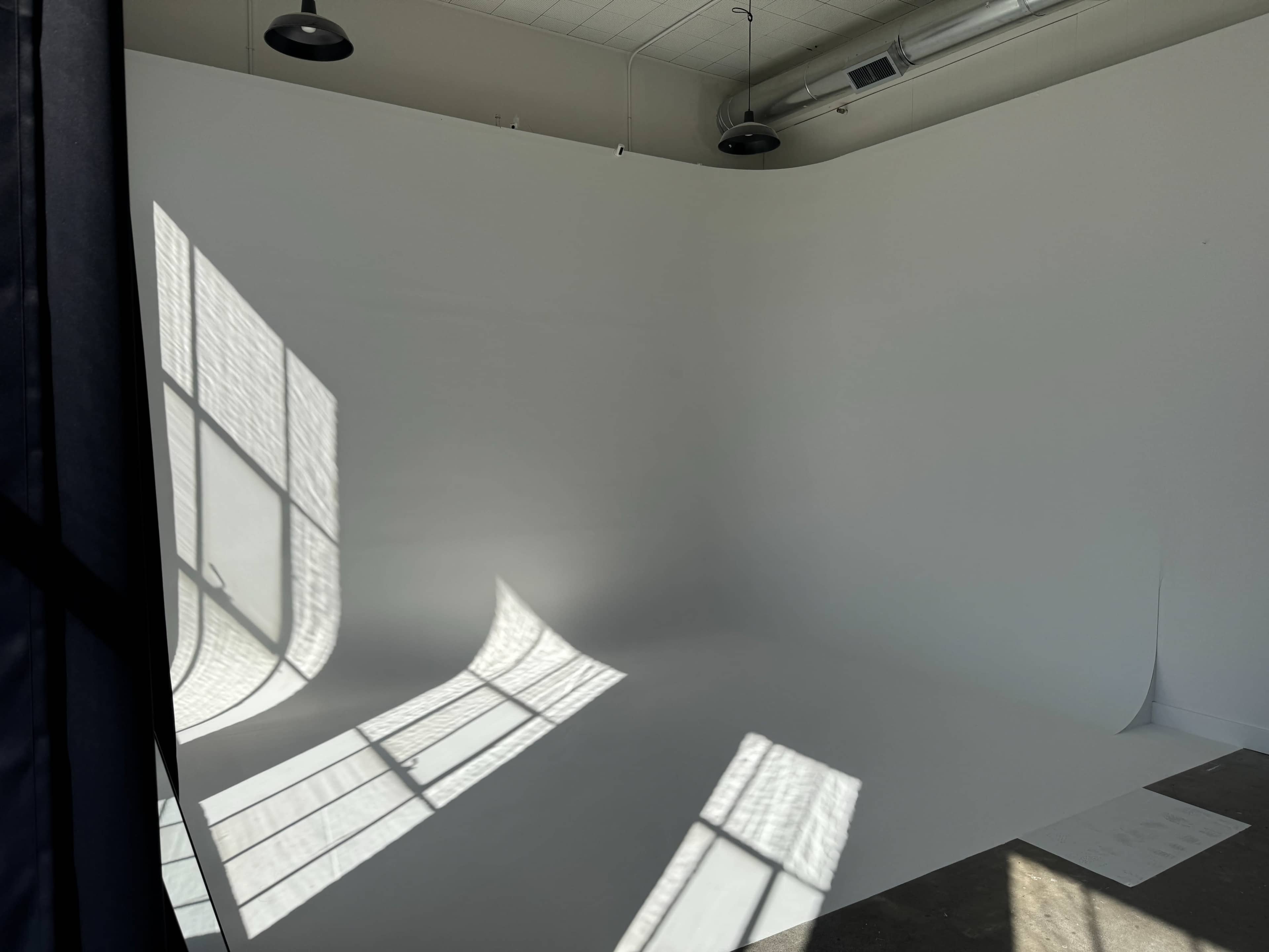 The image shows a bright, empty studio space with a white backdrop and shadows cast by large windows.
