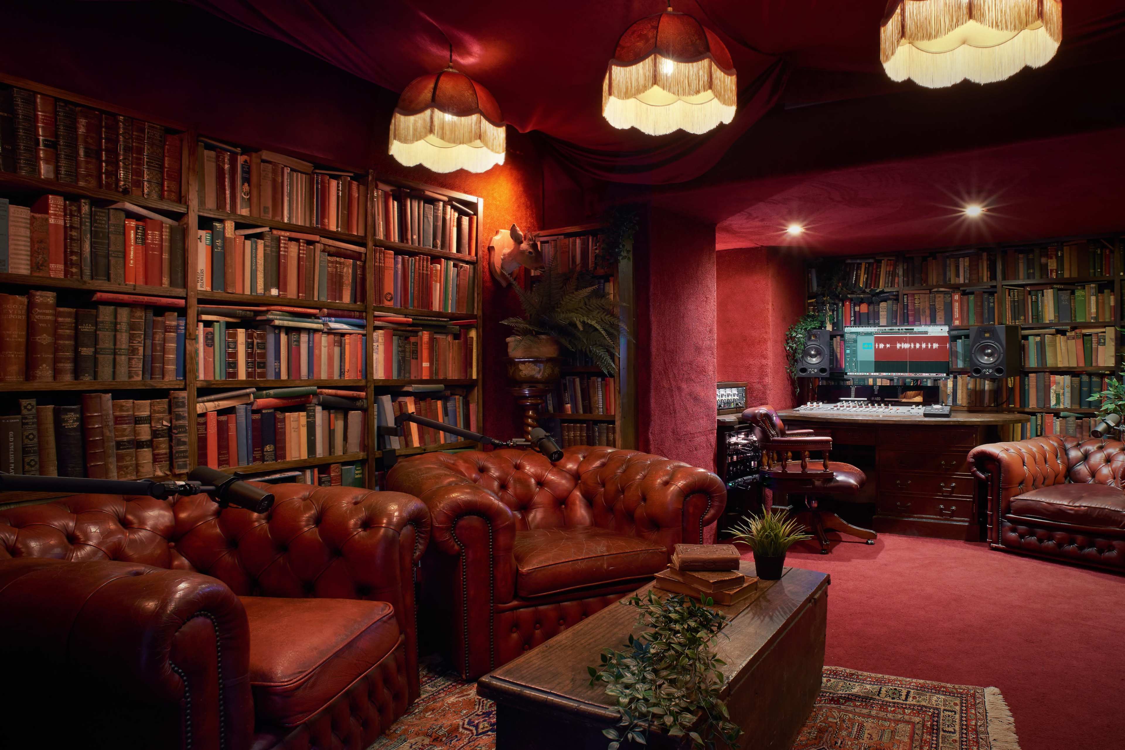 The image shows a cozy, dimly lit room featuring dark red walls, two leather chesterfield sofas, a wooden coffee table, and shelves filled with books, alongside a recording setup equipped with speakers and a computer.