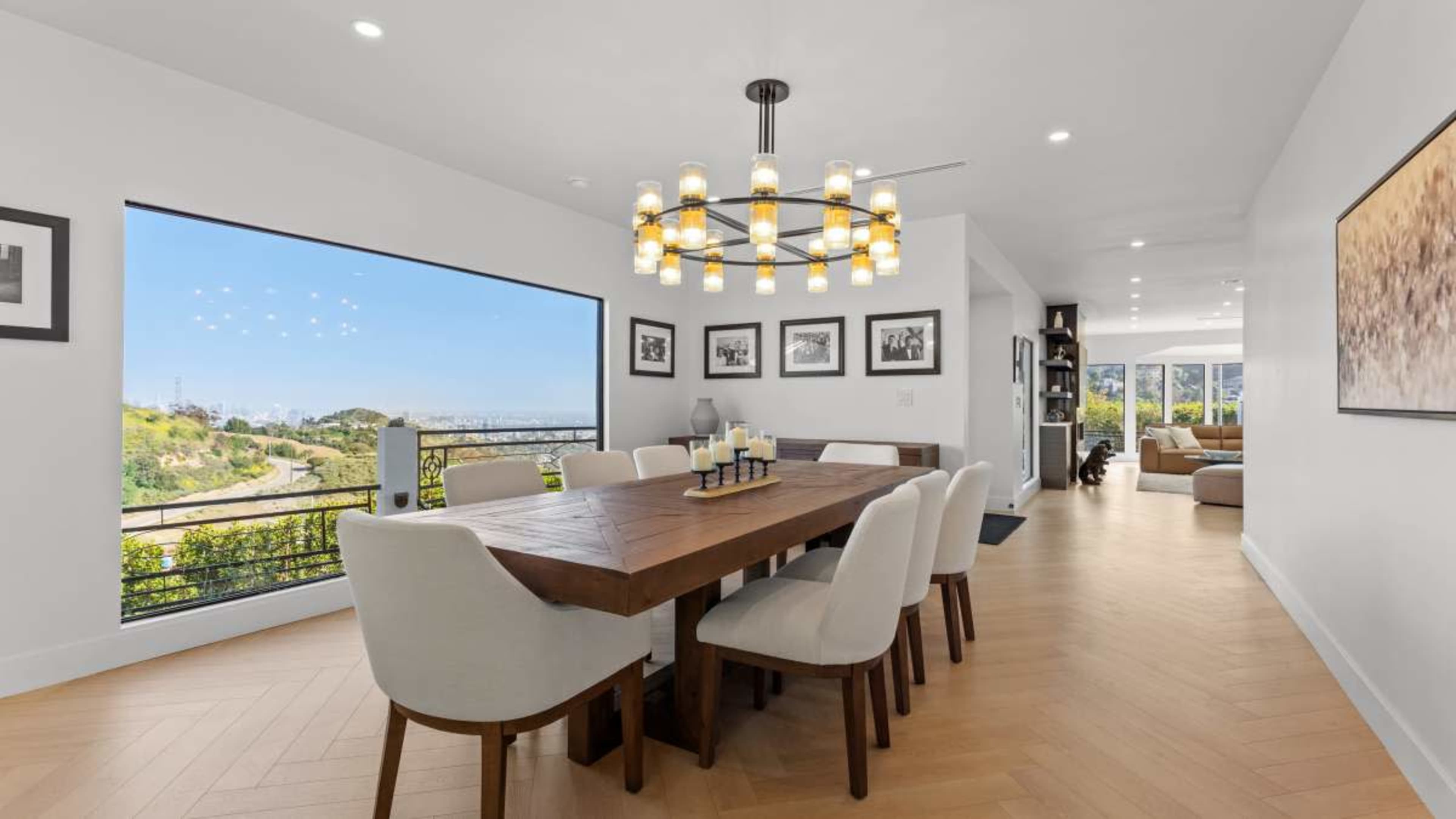 The image shows a modern dining area featuring a large wooden table surrounded by white upholstered chairs, with a view of hills and a cityscape through a large window.