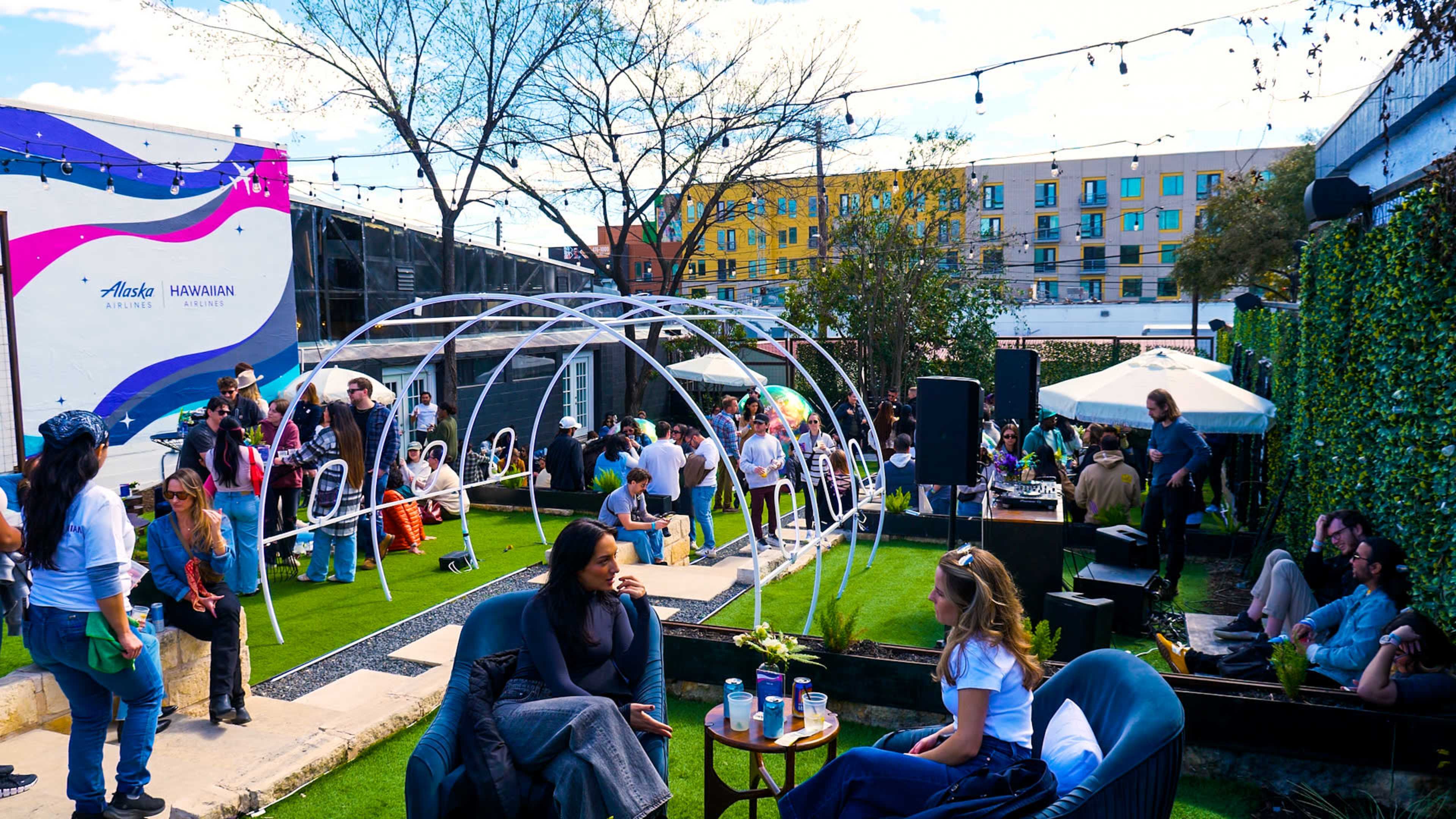 The scene shows a vibrant outdoor event with people socializing, sitting on lounge furniture, and a DJ setup under string lights in a grassy area surrounded by buildings.