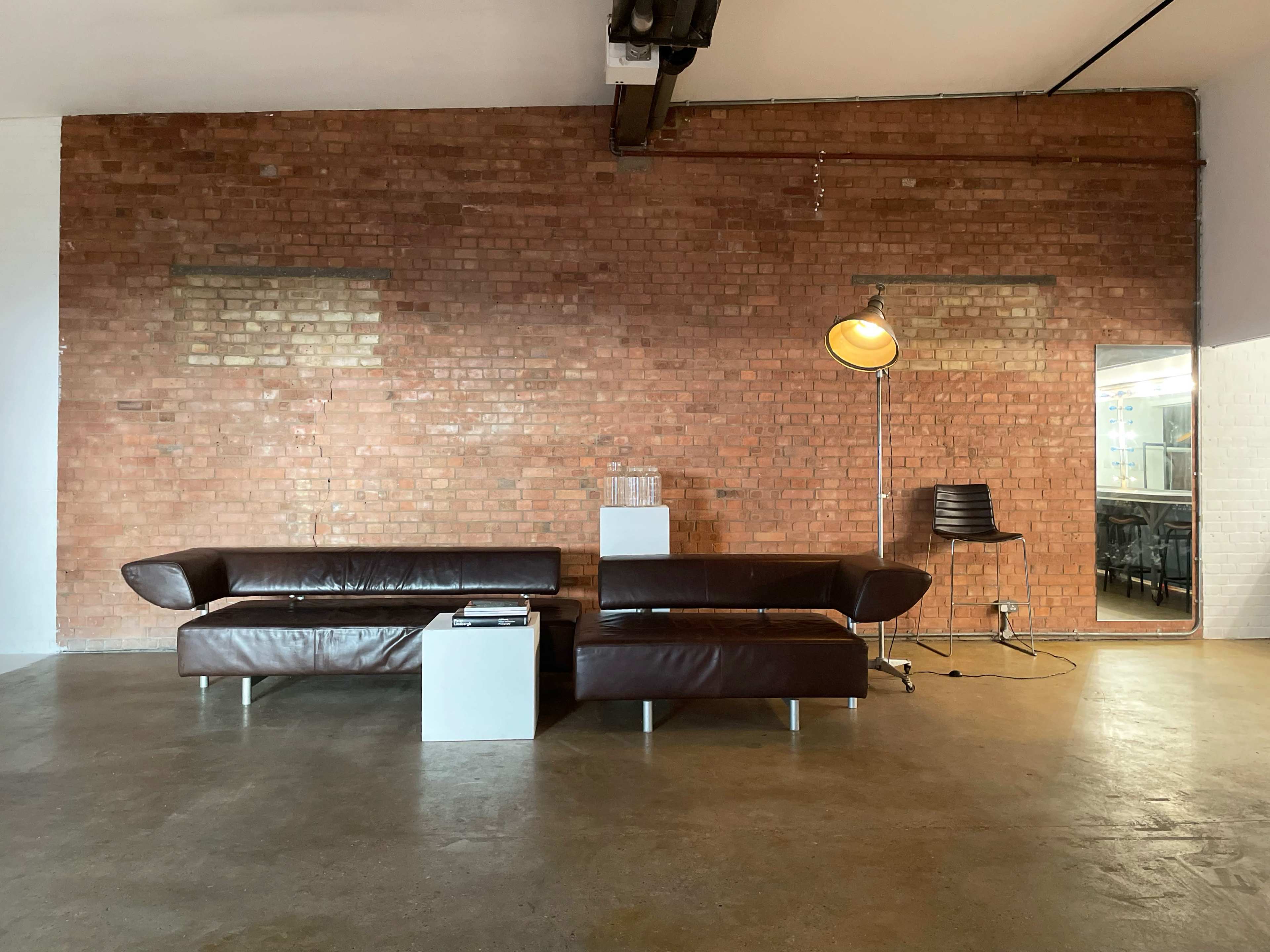 A brown leather sofa and a matching loveseat are positioned against a red brick wall, with a floor lamp and a small white side table nearby.