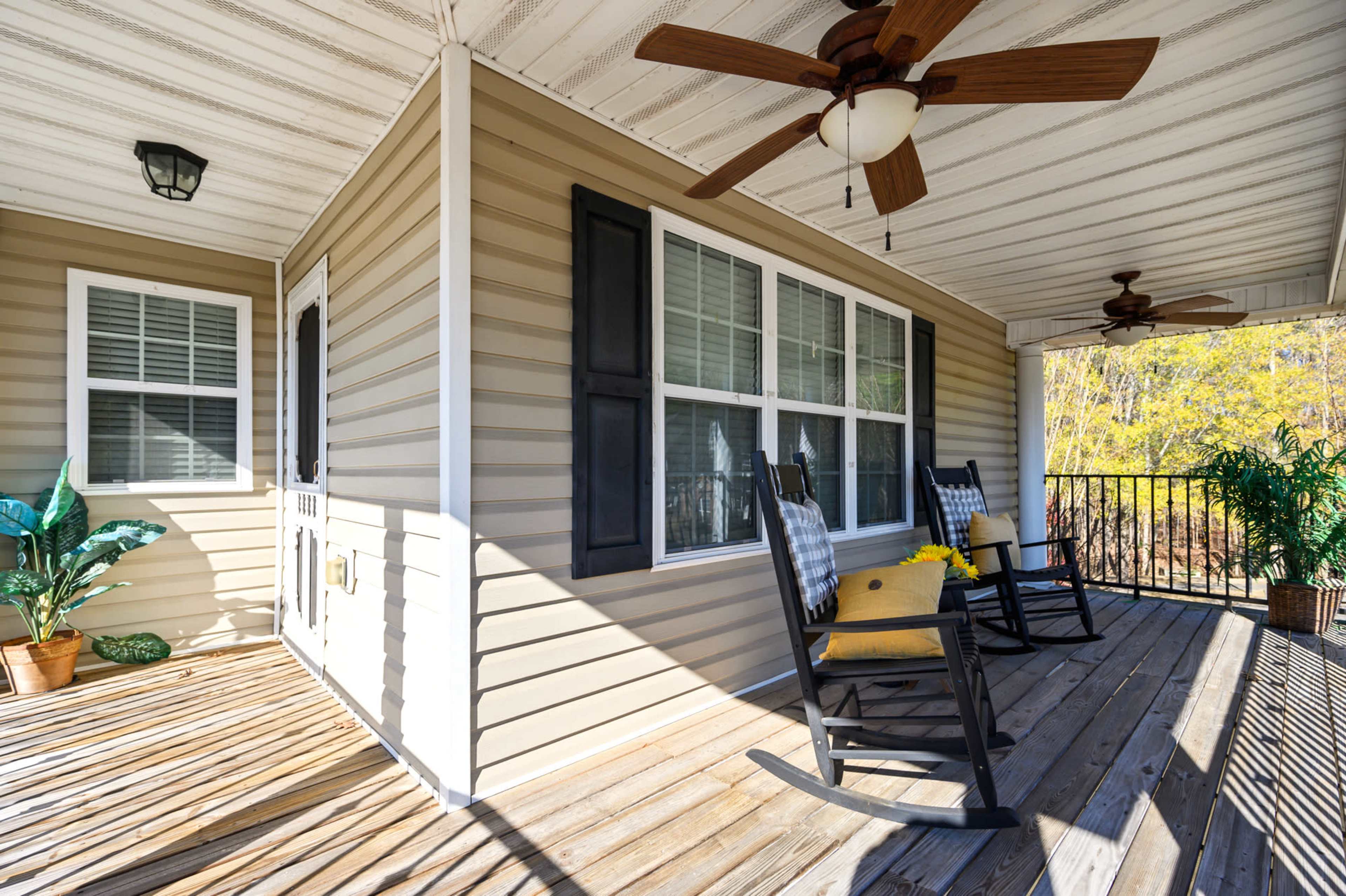 A covered porch featuring two rocking chairs, a houseplant, and large windows, with wooden decking and two ceiling fans overhead.