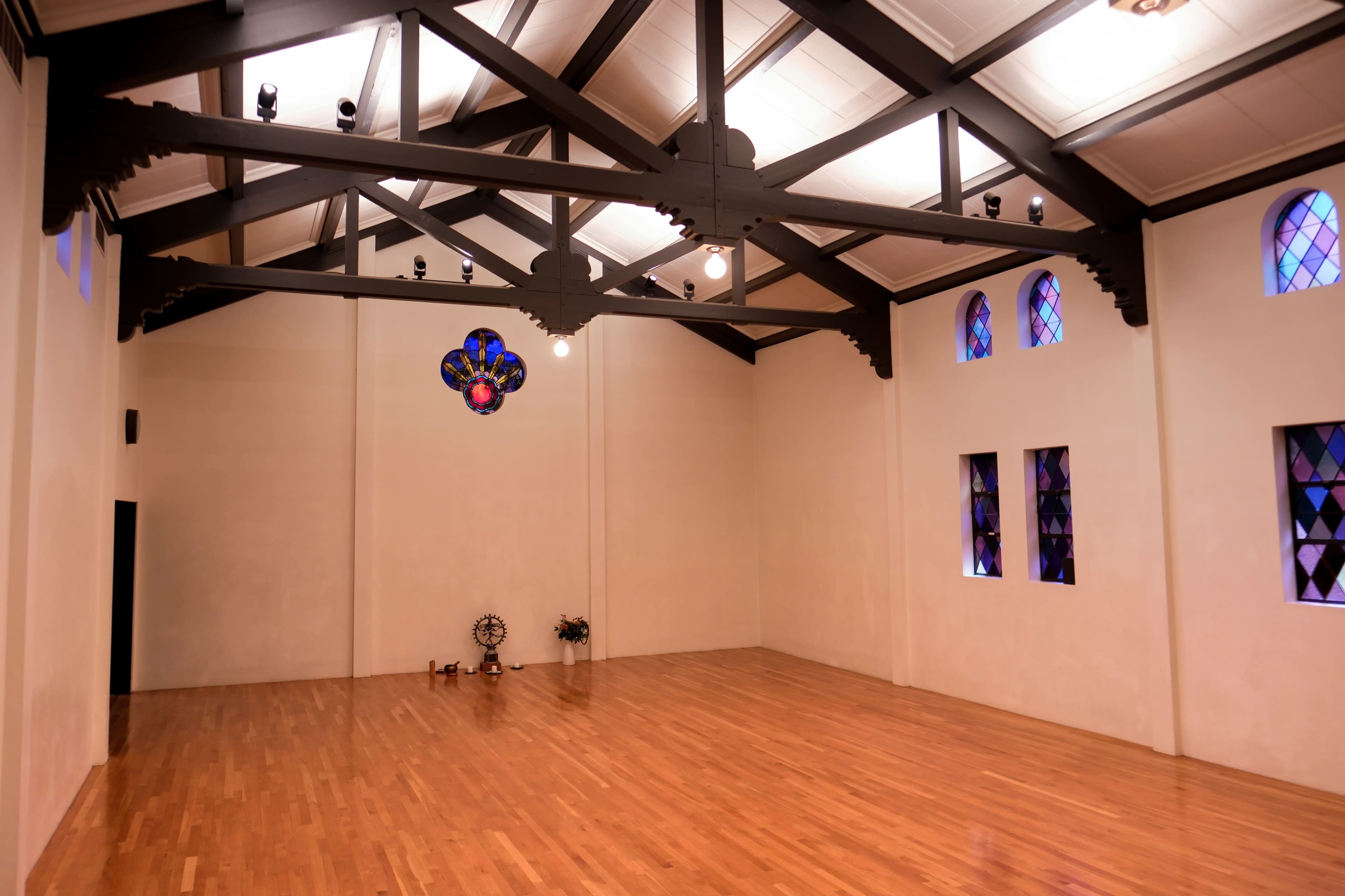 The image shows a spacious, empty room with wooden flooring, high ceilings supported by black beams, and stained glass windows.