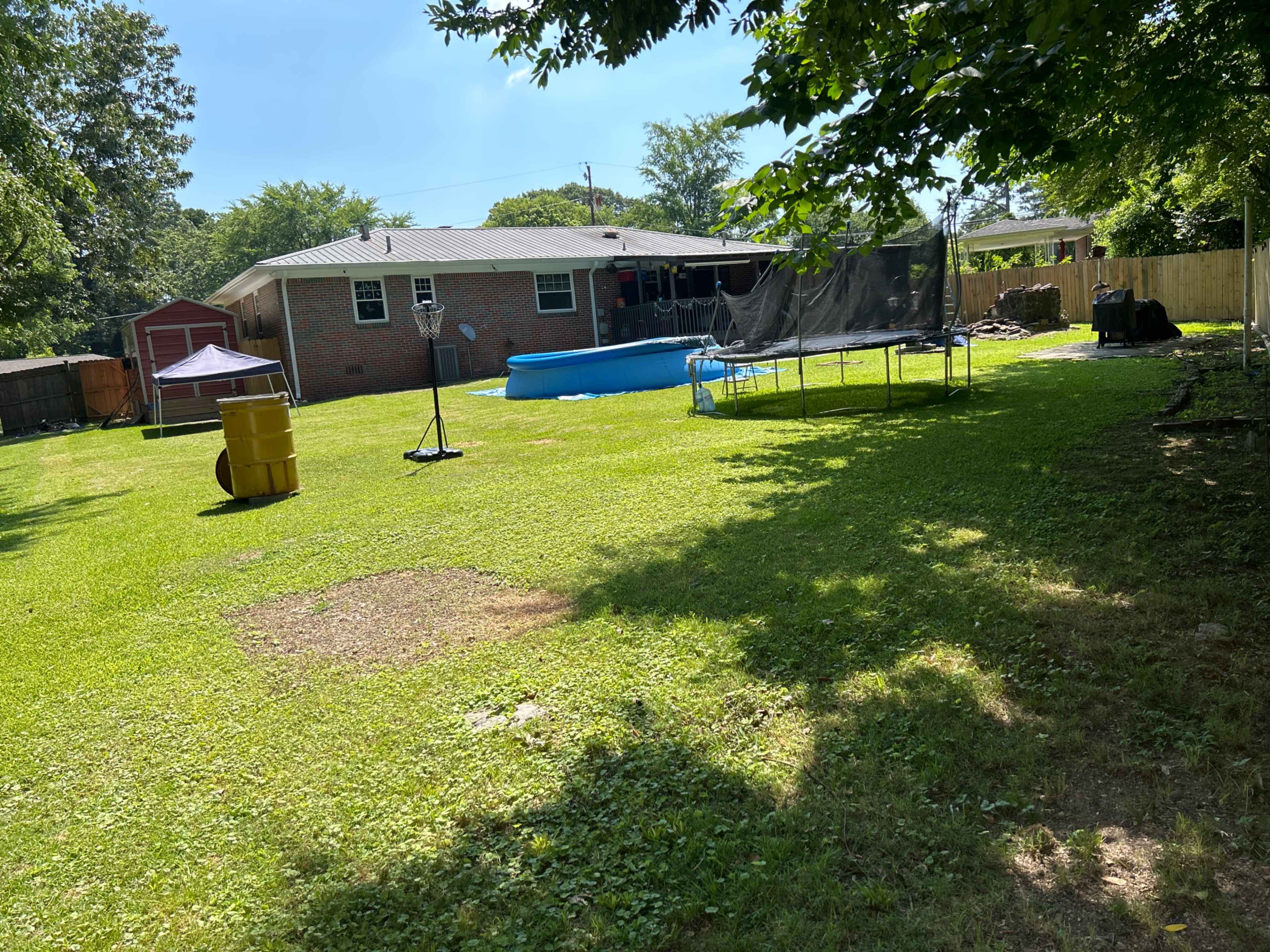 A backyard featuring a swimming pool, a trampoline, a yellow barrel, and a brick house in the background.