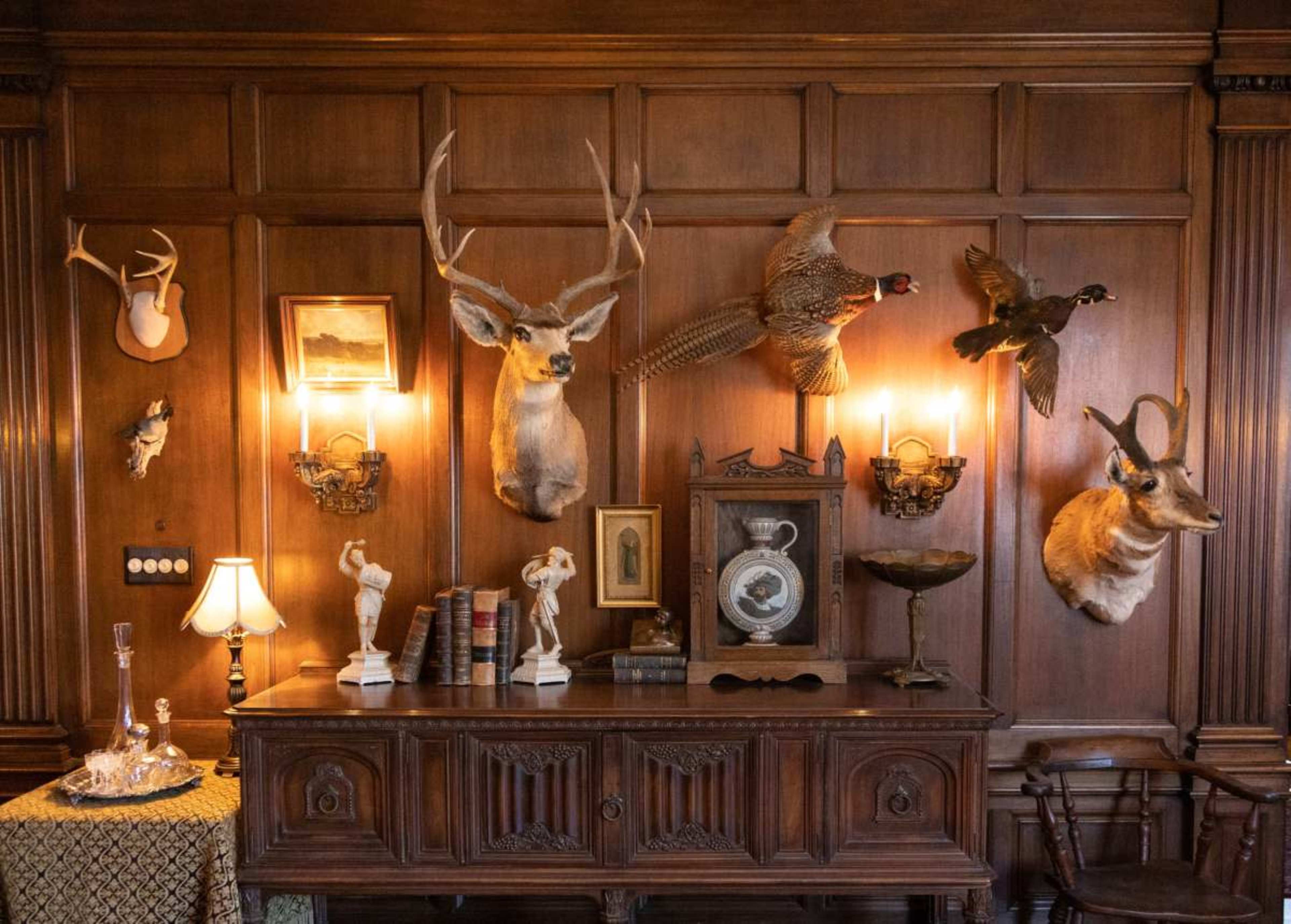 The image shows a wooden wall adorned with mounted animal heads and a collection of vintage items, including books and a clock, displayed on a sideboard beneath two wall sconces.