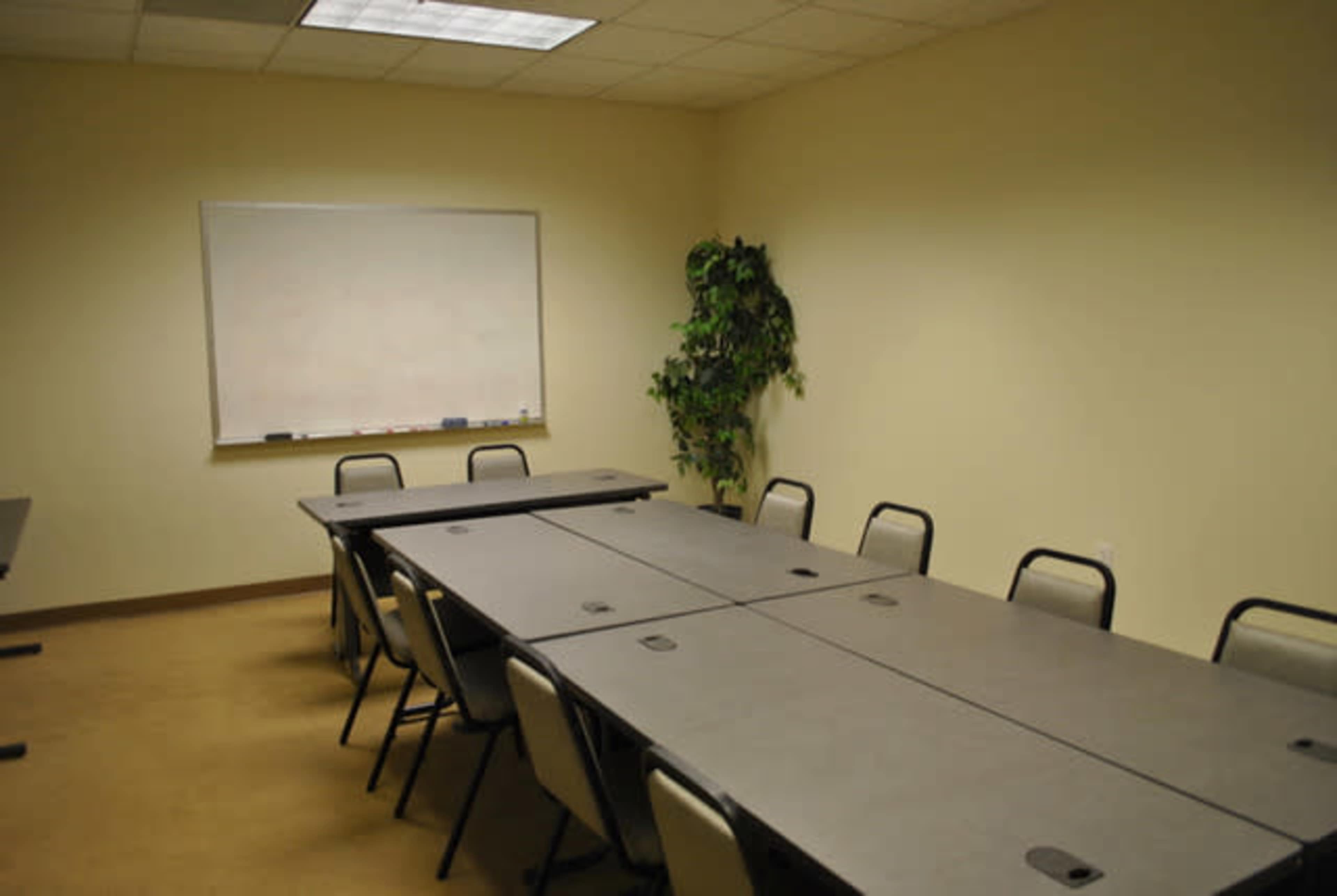A sparse conference room features several rectangular tables arranged in a U-shape, with a whiteboard and a plant in the corner.