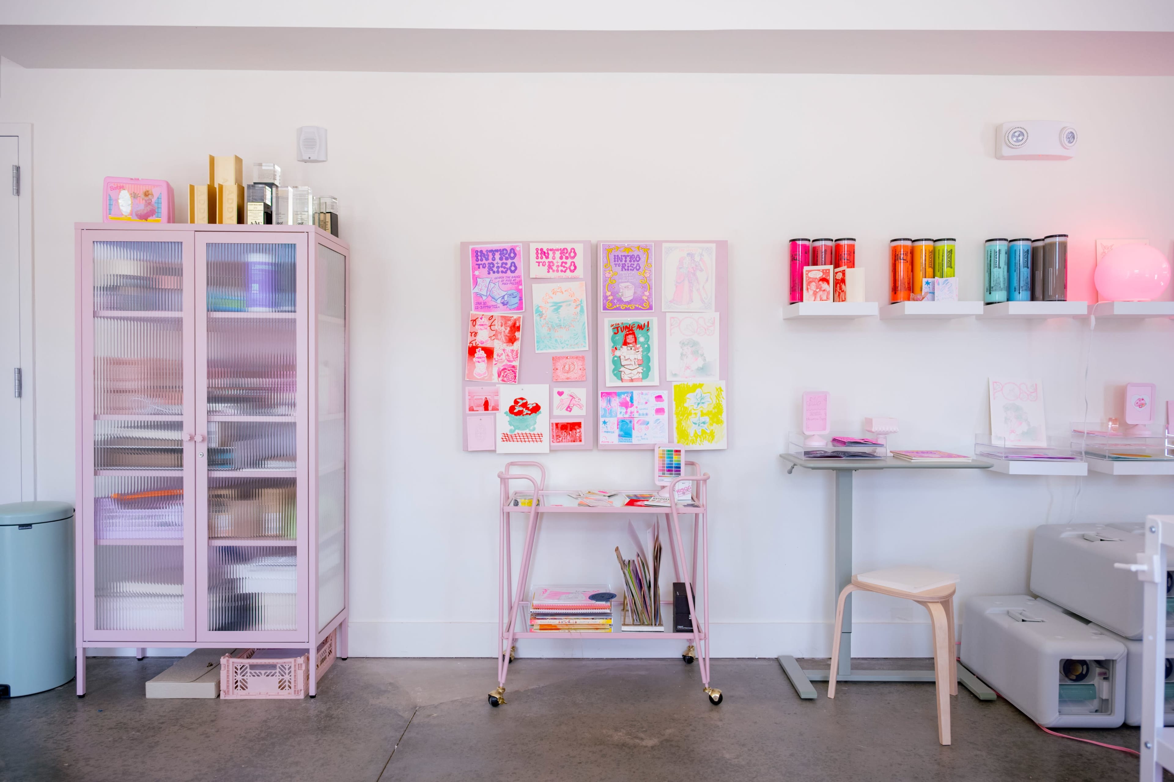 The image shows a bright, pastel-themed workspace featuring a pink cabinet, a bulletin board with colorful artwork, and a table with various art supplies.
