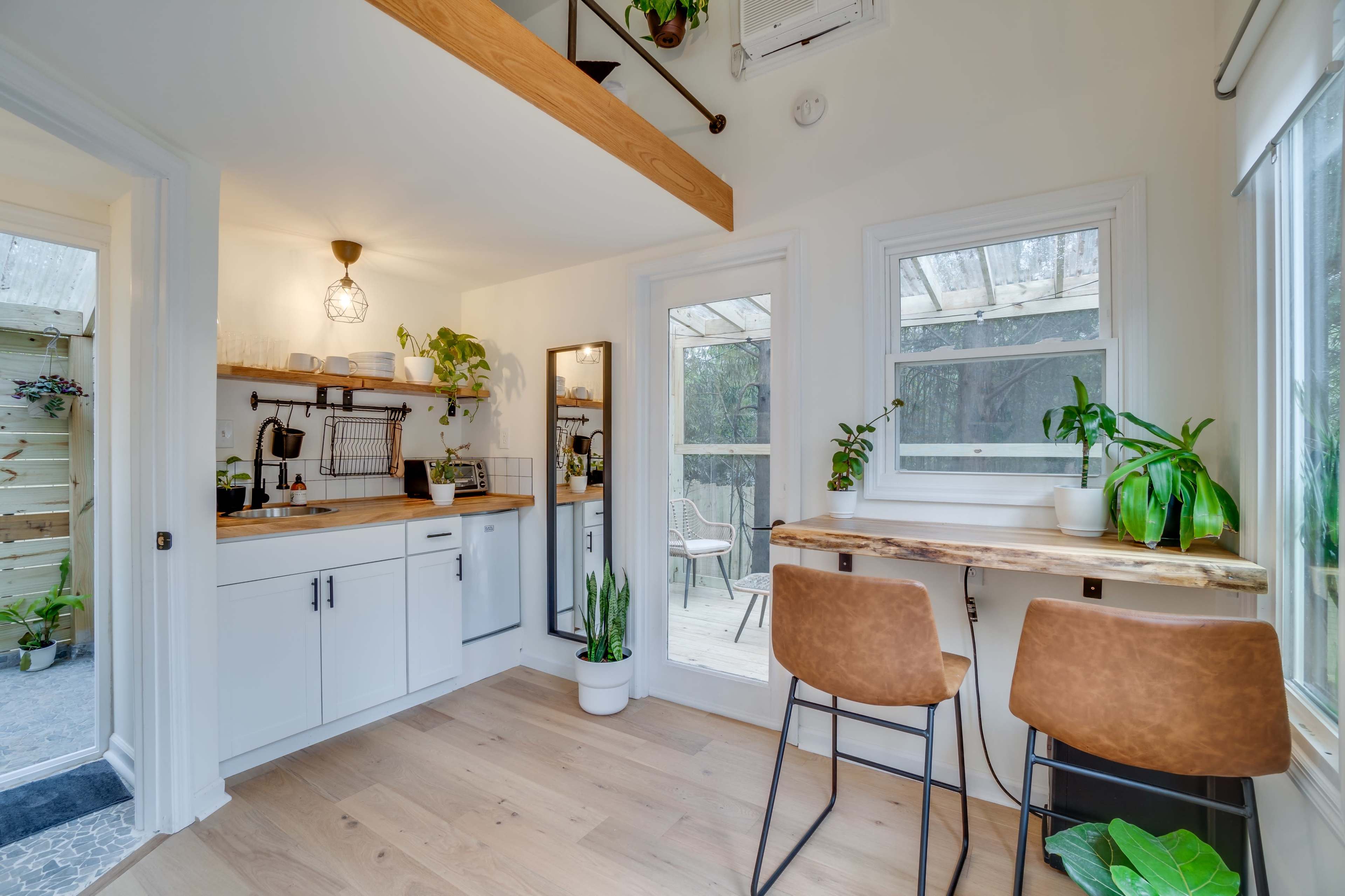 A small, modern kitchen area with white cabinetry, a wooden countertop, and a seating space featuring two brown chairs next to a large window with plants.