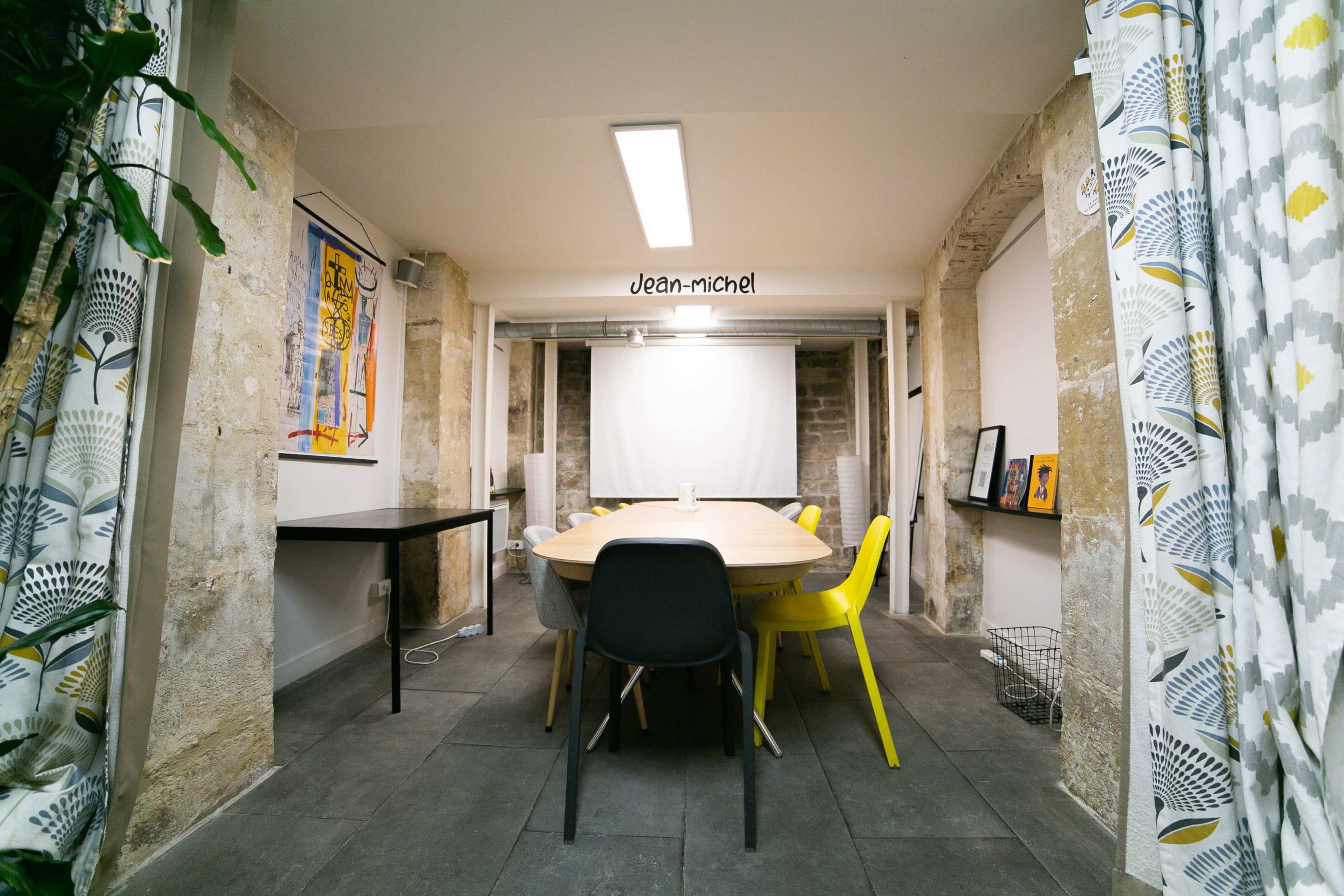 A modern meeting room features a large circular table surrounded by colorful chairs, with a projector screen at the front and stone walls.