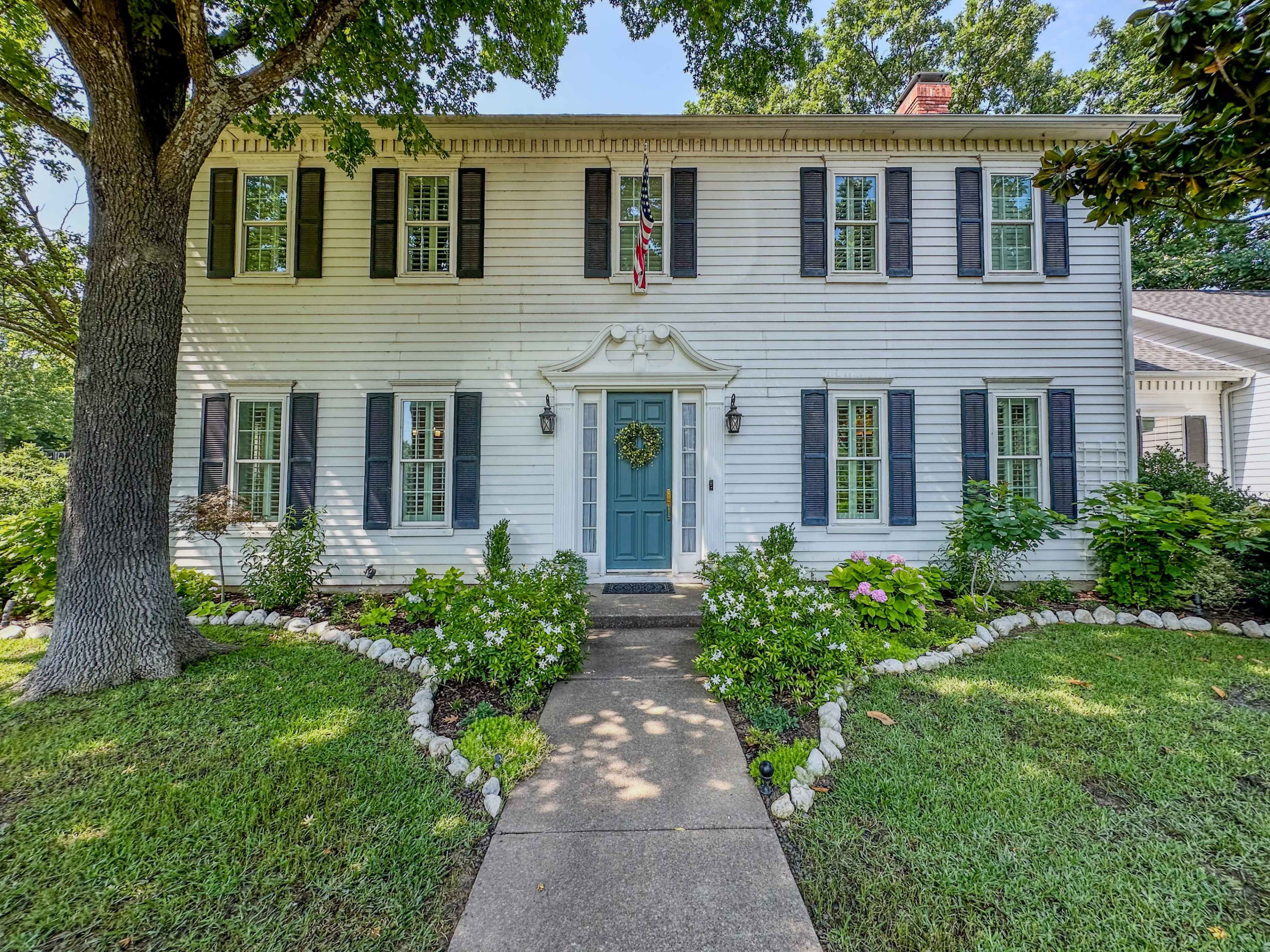 The image depicts a two-story white house with black shutters, a central blue door, and a well-maintained garden pathway lined with stones.